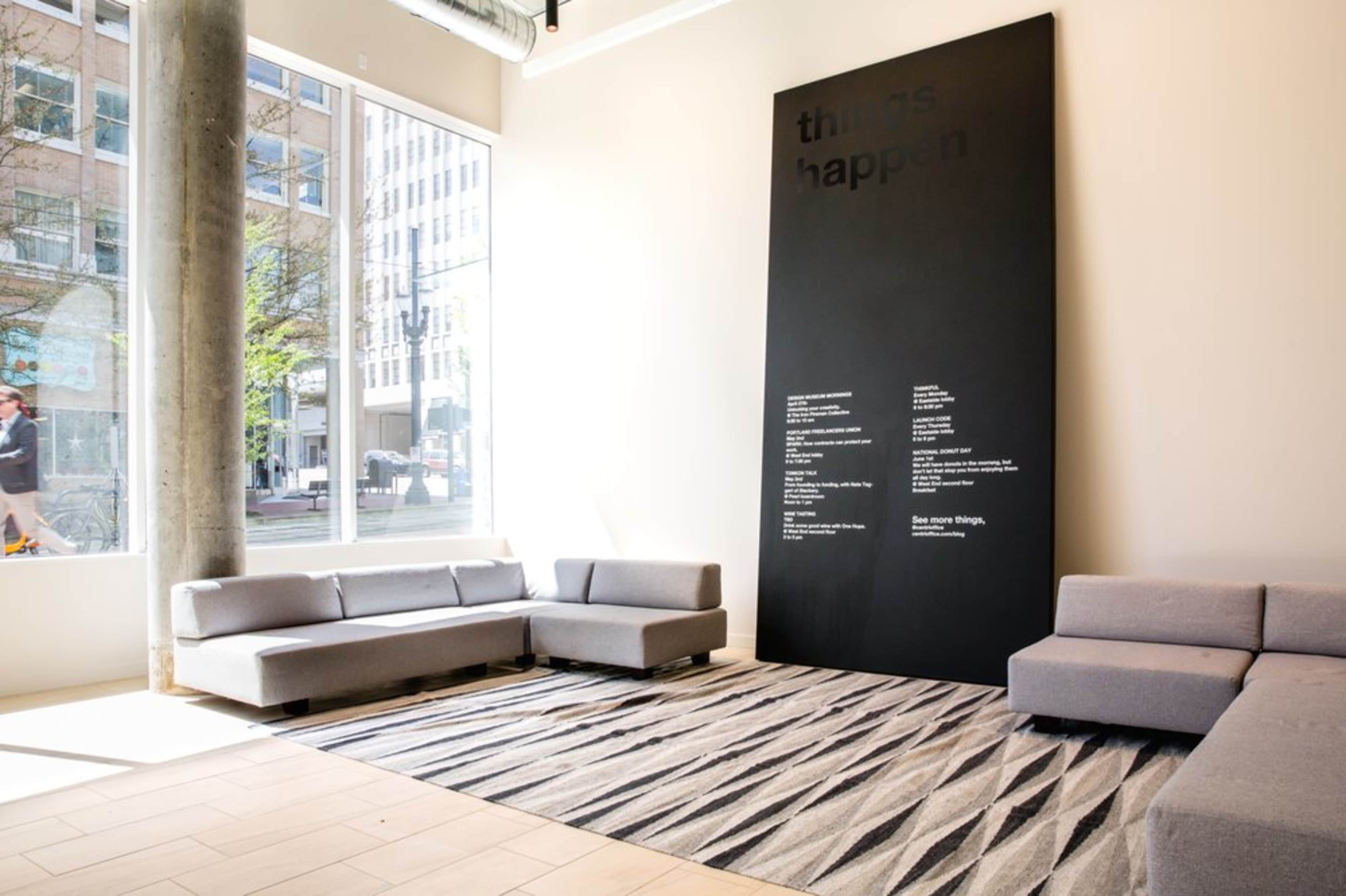 A spacious interior features gray couches arranged around a geometric rug, with a large black wall displaying white text.