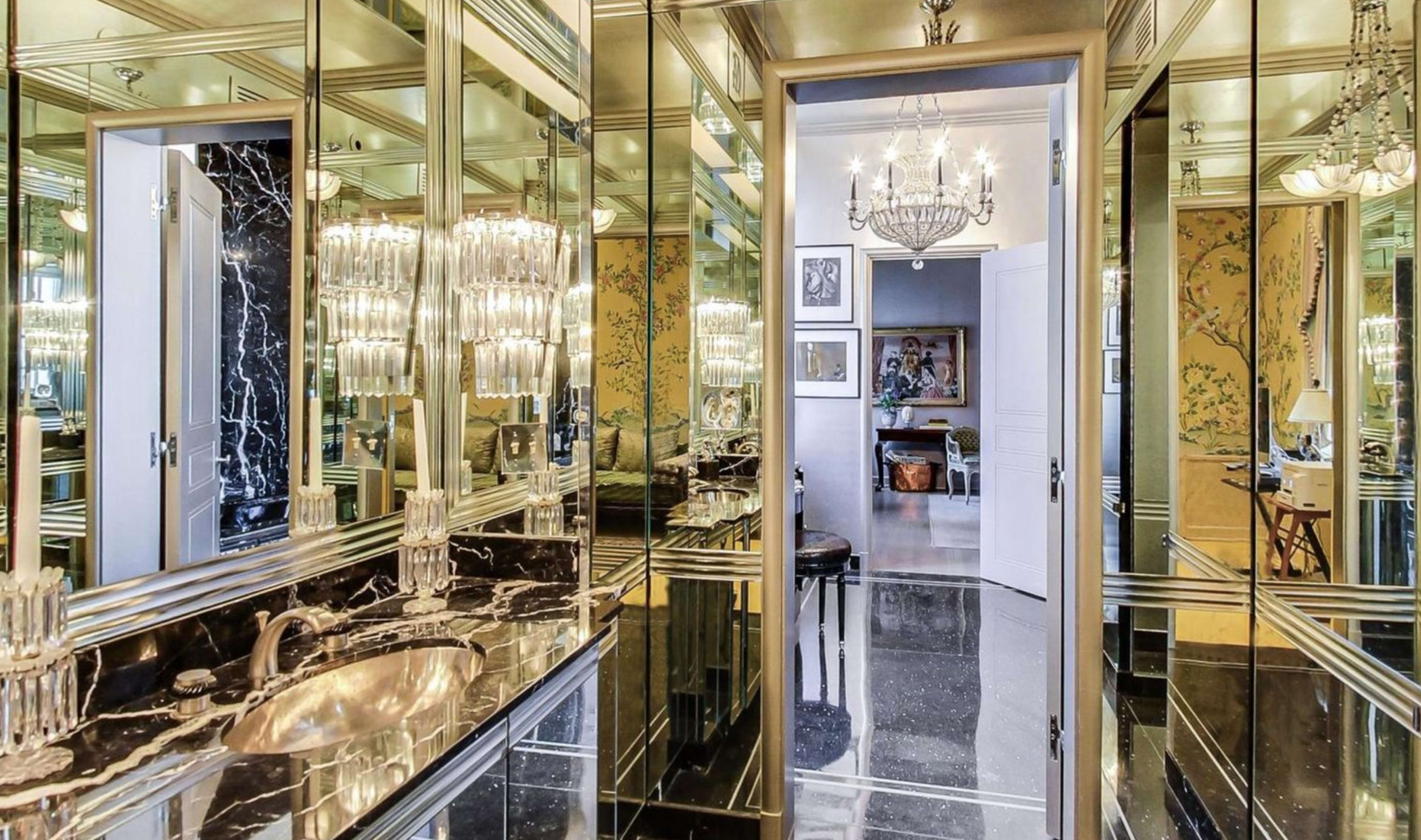 A luxurious bathroom with mirrored walls, a black marble vanity, and a chandelier, leading into a well-decorated adjoining room.
