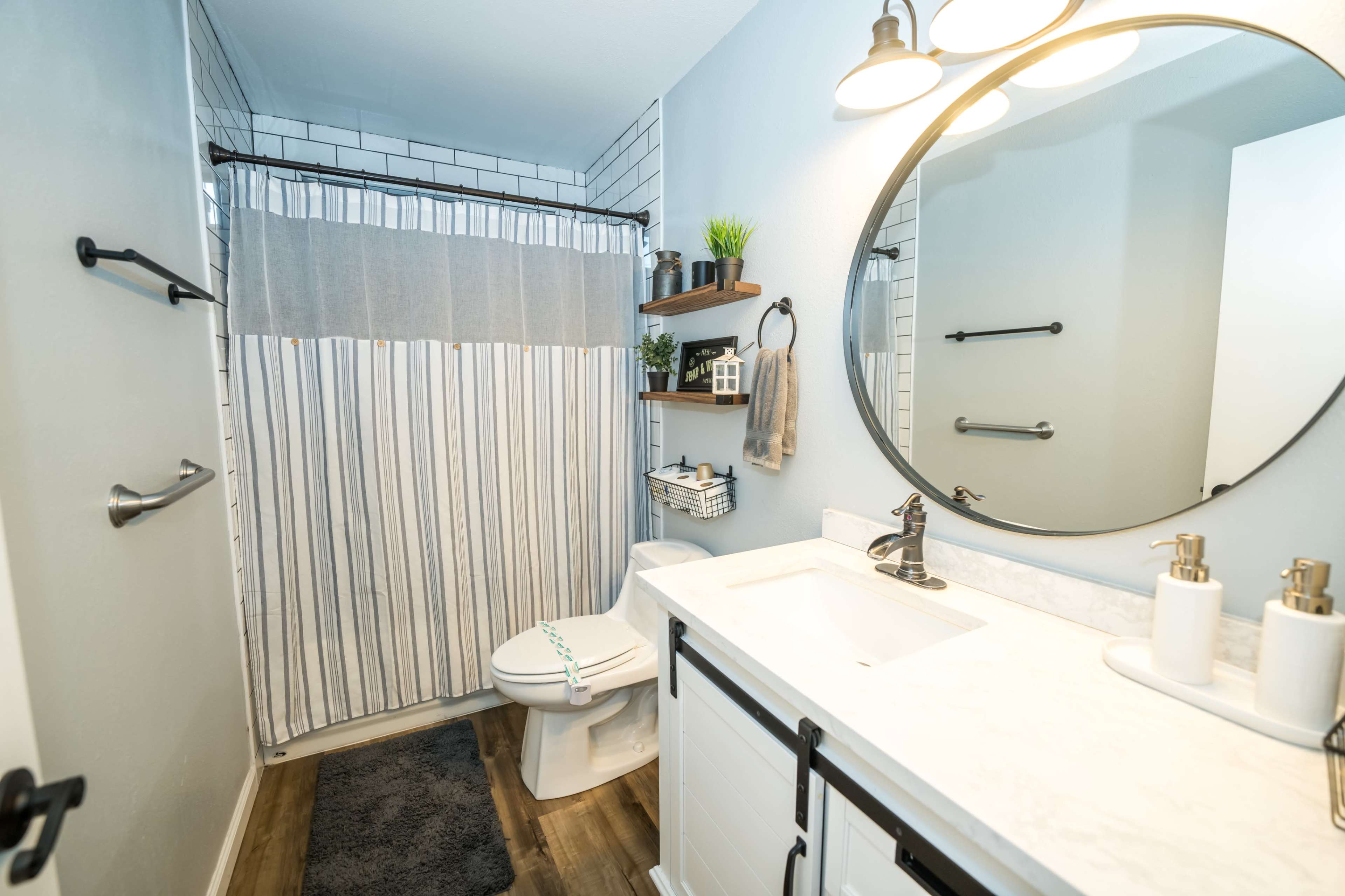 A modern bathroom featuring a shower with a striped curtain, a white countertop with two sinks, a round mirror, and shelves with decorative items.