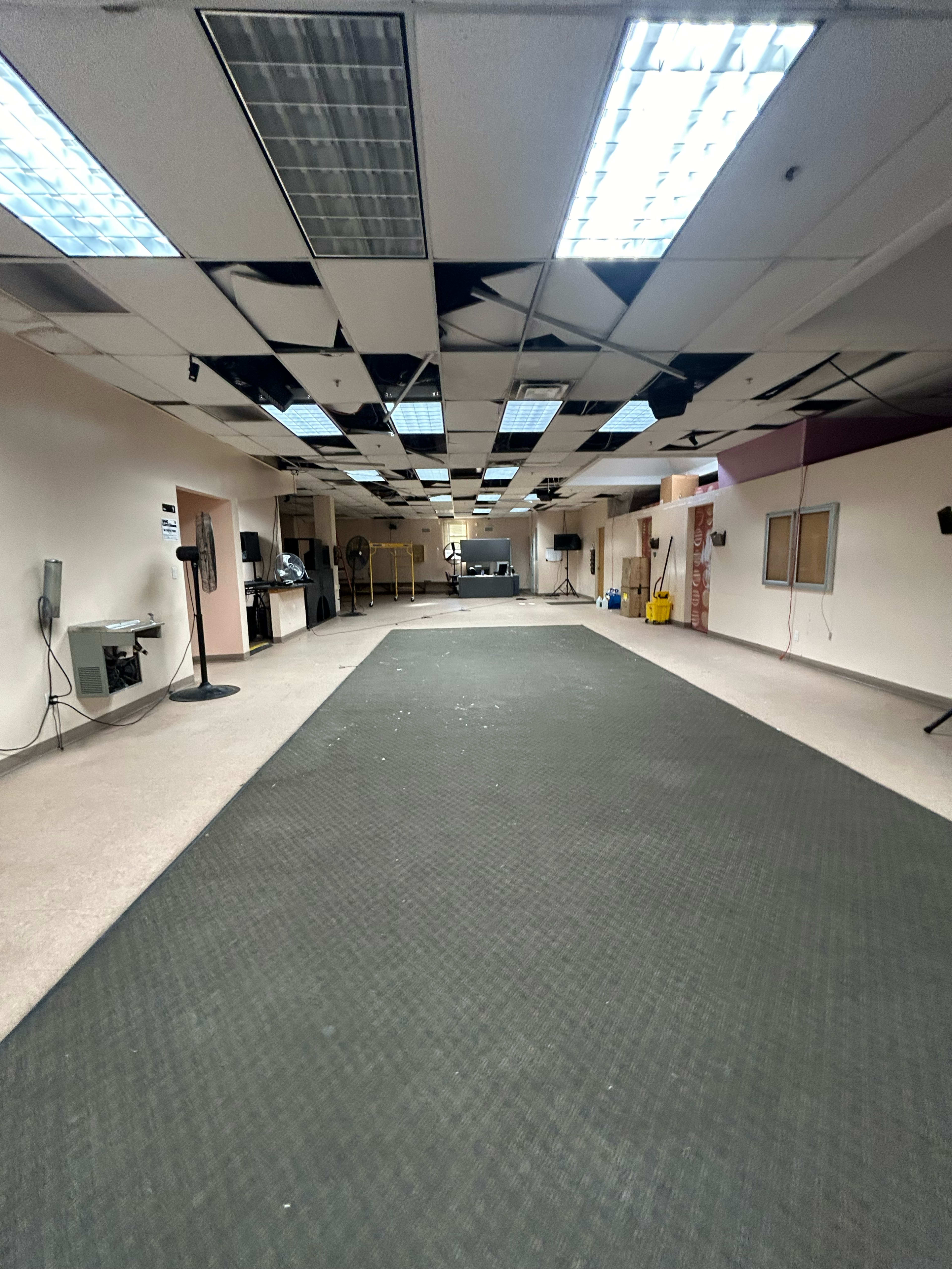 The image shows an empty, wide room with a dark carpet, overhead lighting, and visible equipment along the walls.