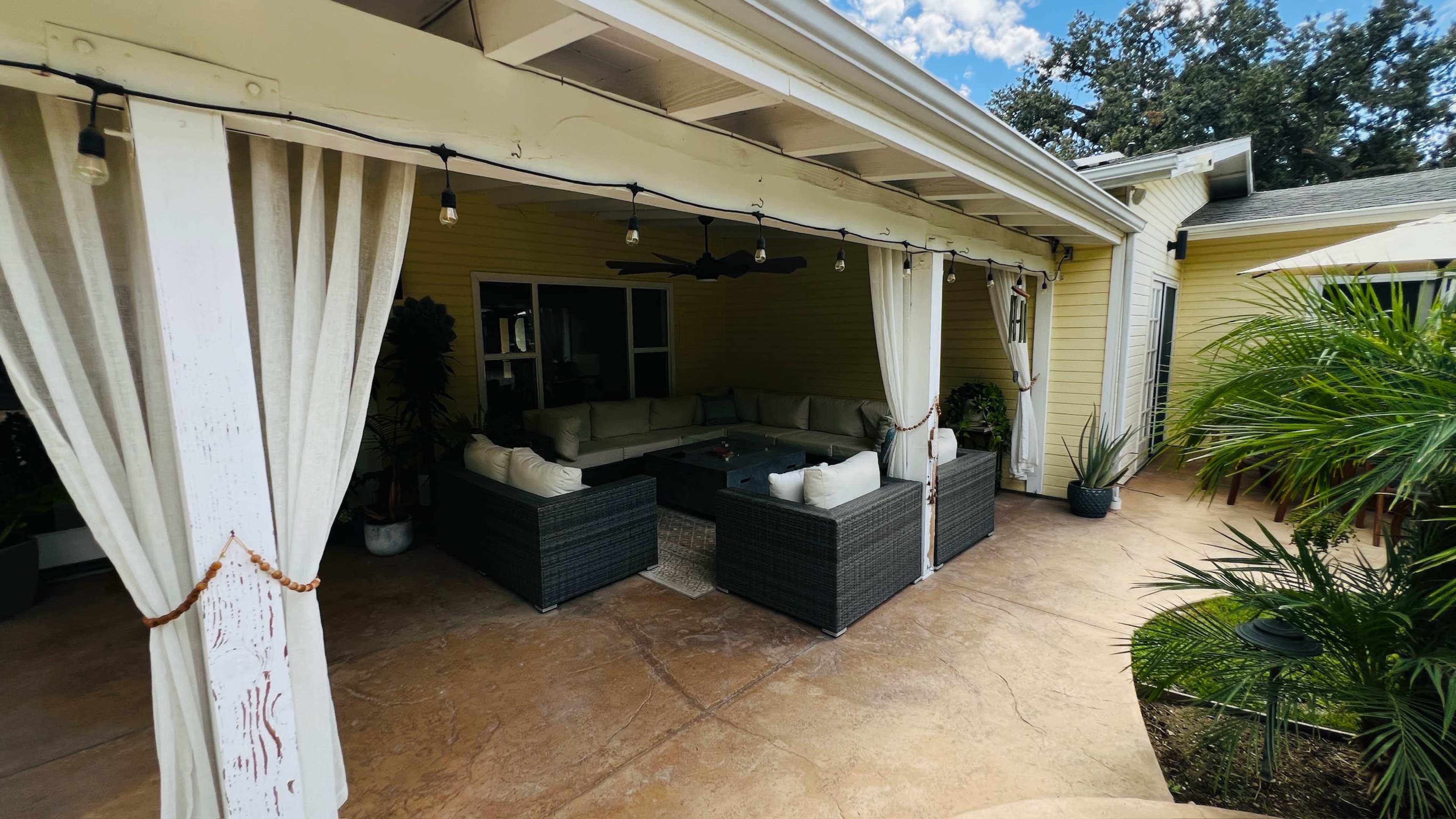 The image shows a covered outdoor patio area featuring a sectional seating arrangement, a coffee table, and decorative plants.
