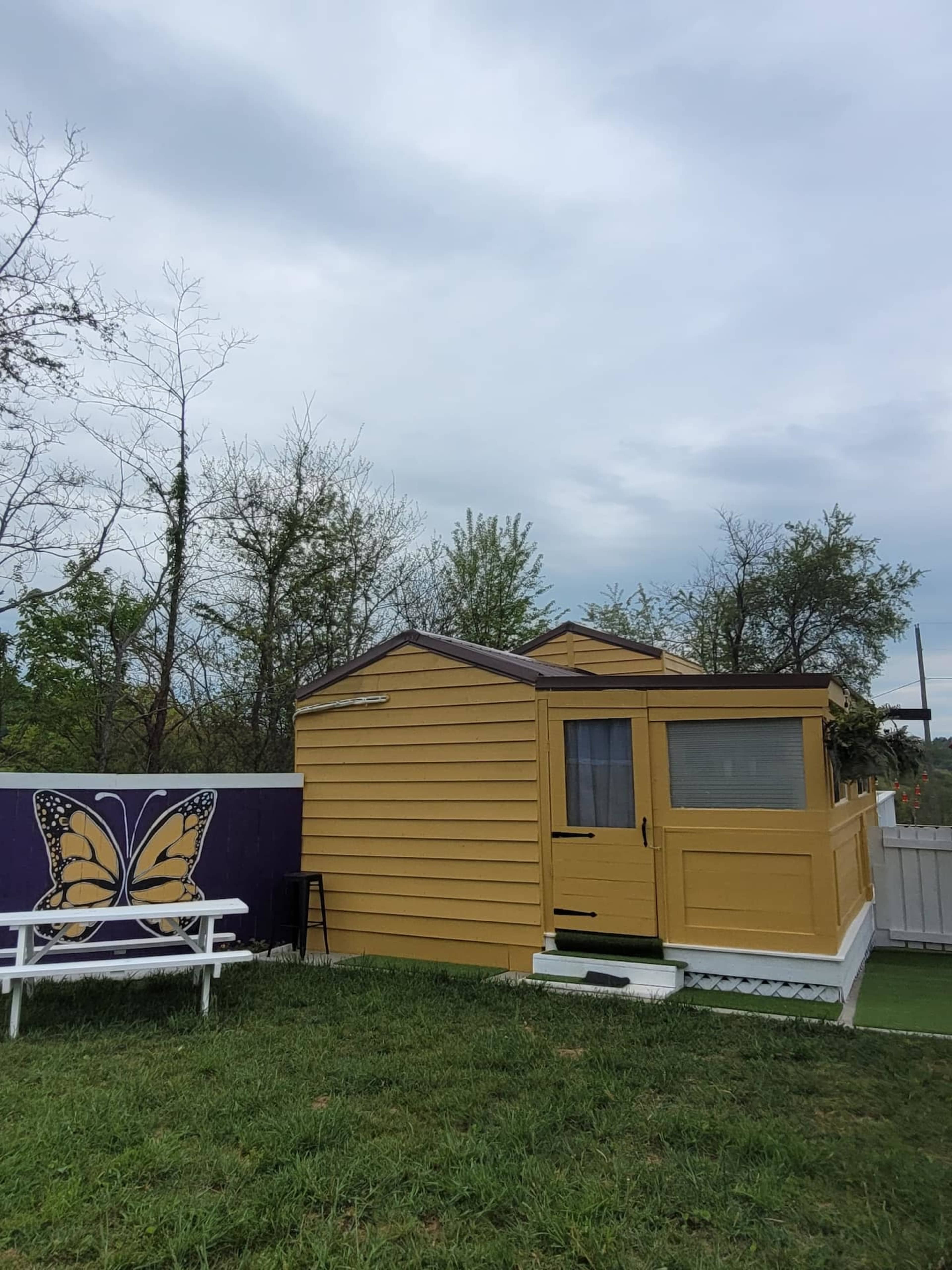 A small yellow house with a porch is situated next to a purple wall featuring a butterfly mural, surrounded by grass and trees under a cloudy sky.