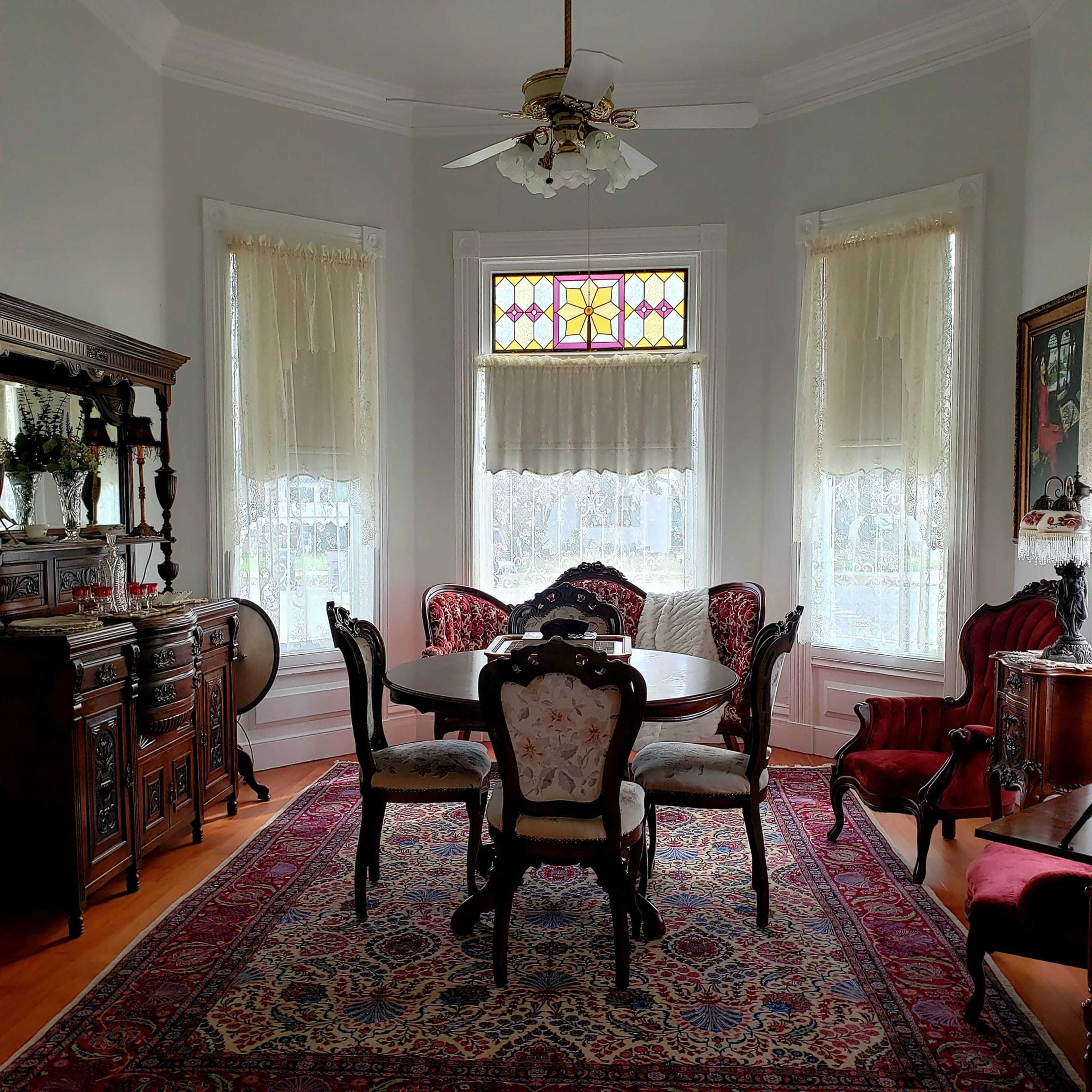 The image shows a vintage dining room with a round table, upholstered chairs, and ornate wooden furniture, illuminated by natural light through stained glass windows.