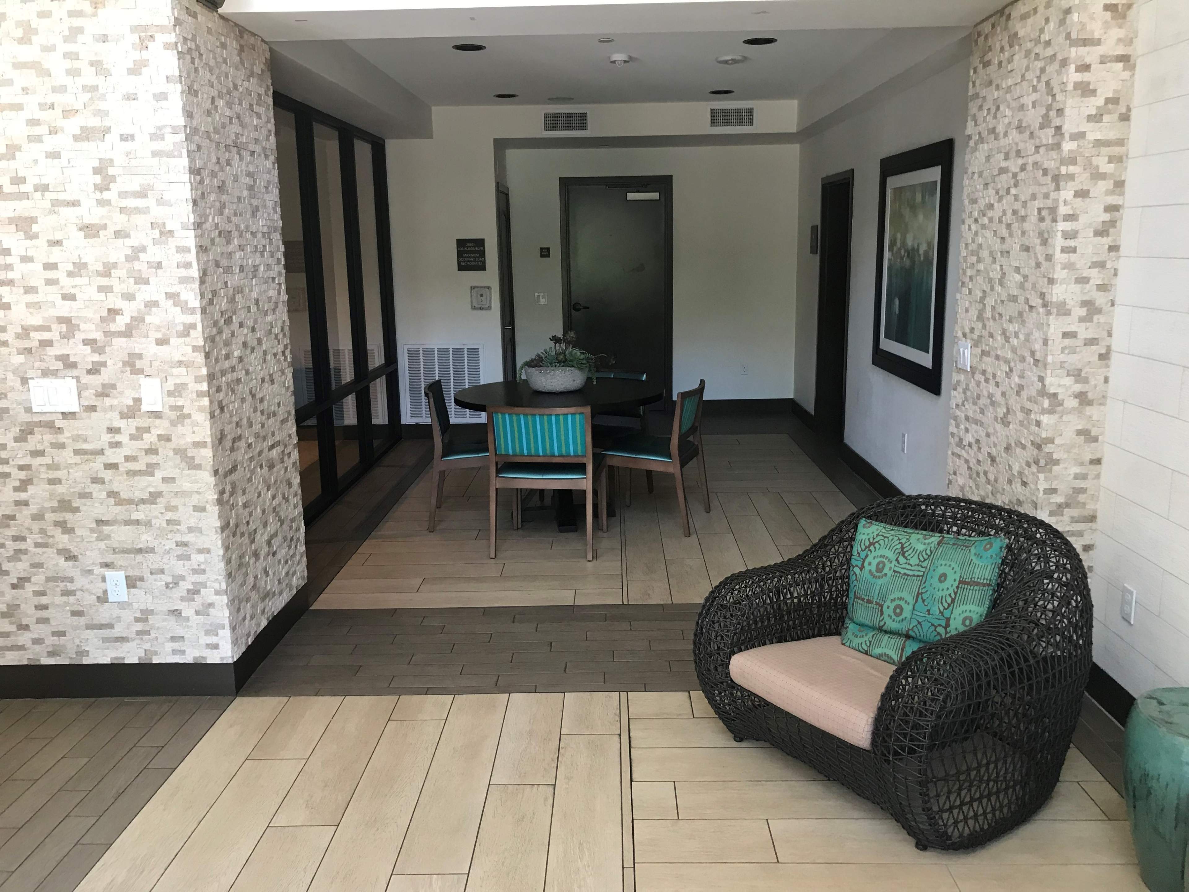 A modern lounge area features a round table with teal chairs and a woven armchair in a bright, open space with decorative wall tiles.
