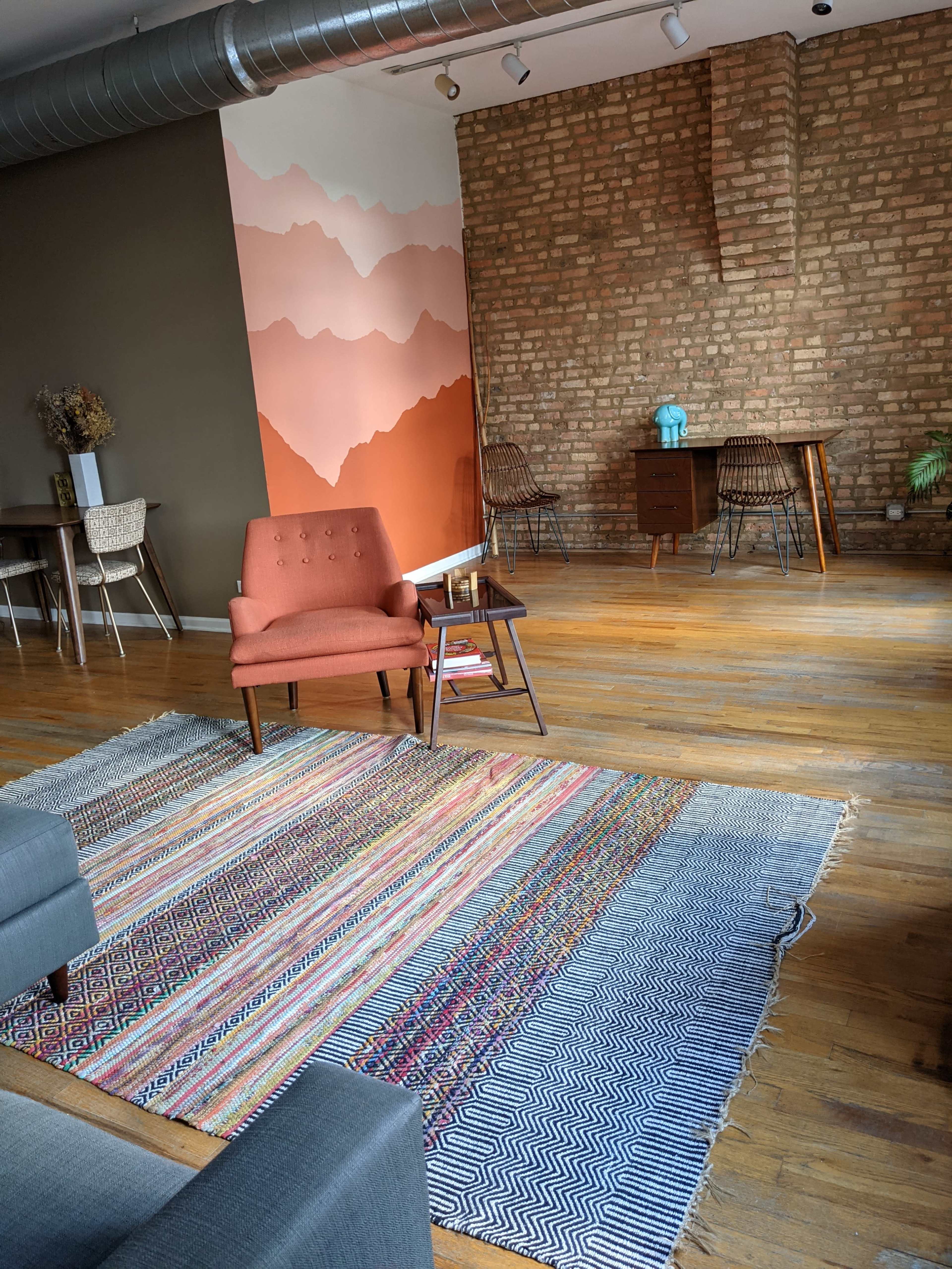 The image shows a modern interior with a patterned rug, an orange armchair, a wooden table, and a mural of mountains on the wall.
