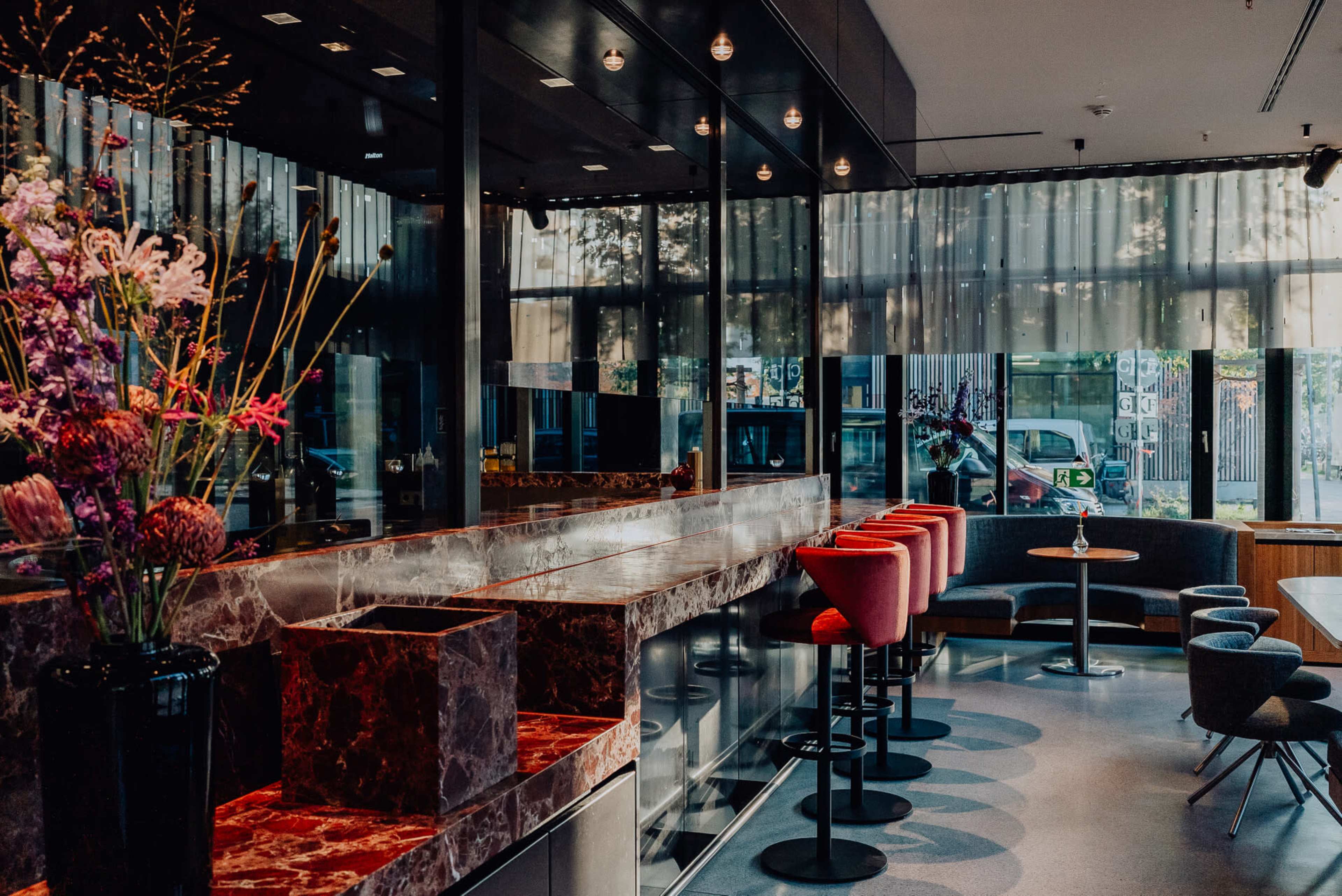 The image shows a modern café interior featuring a marble countertop, red bar stools, and large windows that let in natural light.