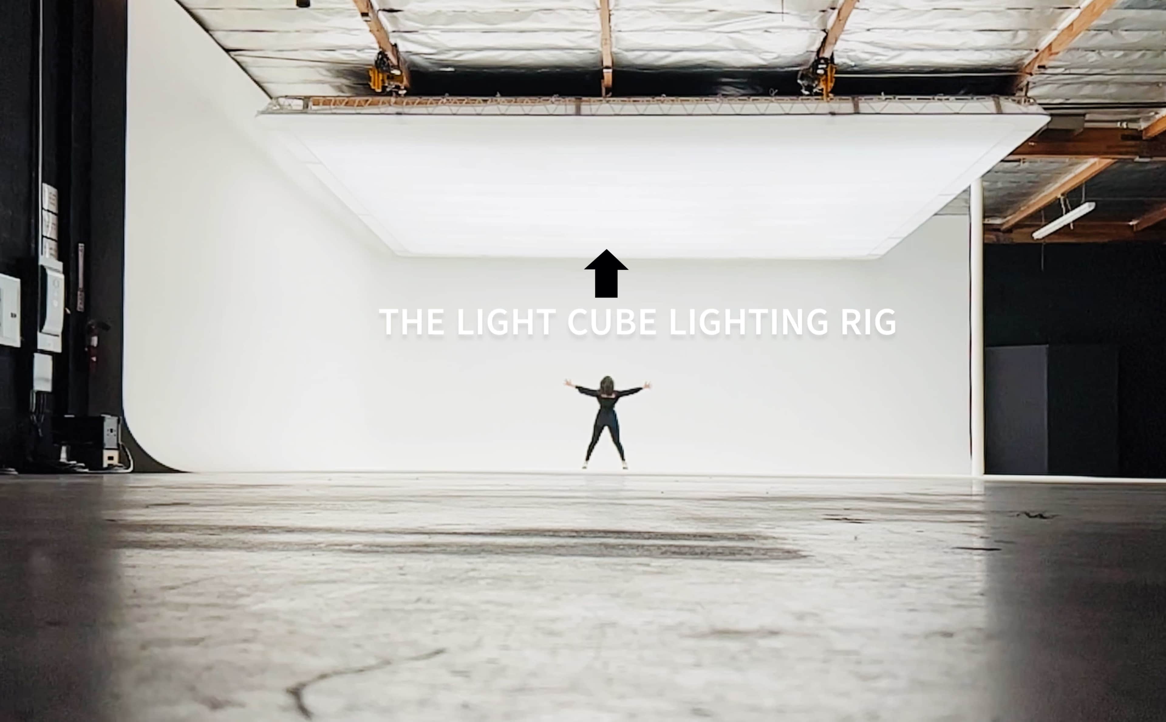 A person stands with arms outstretched in a spacious studio featuring a large overhead lighting rig.