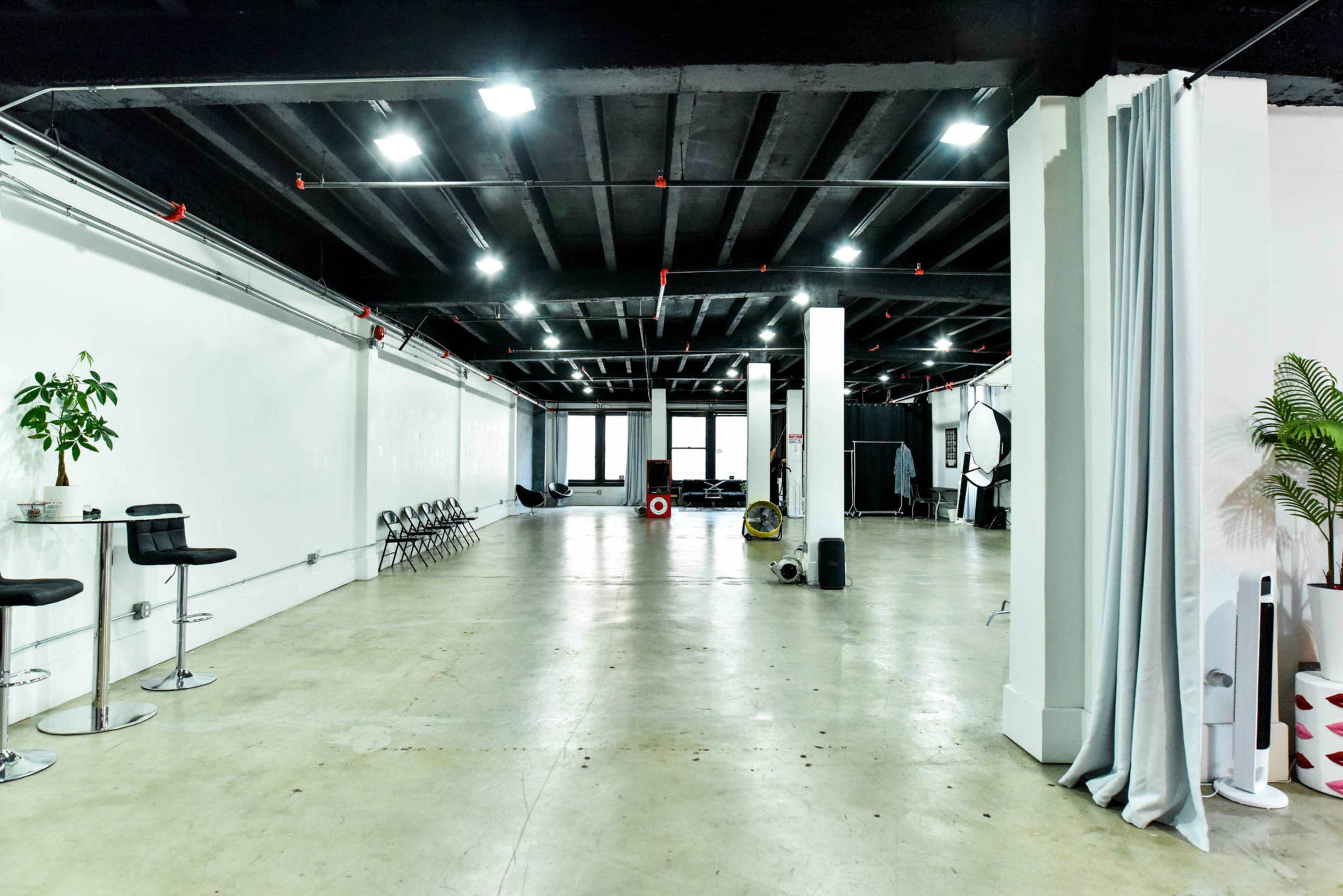 The image shows a spacious, minimalistic studio with concrete flooring, white walls, and a few chairs and plants scattered throughout.