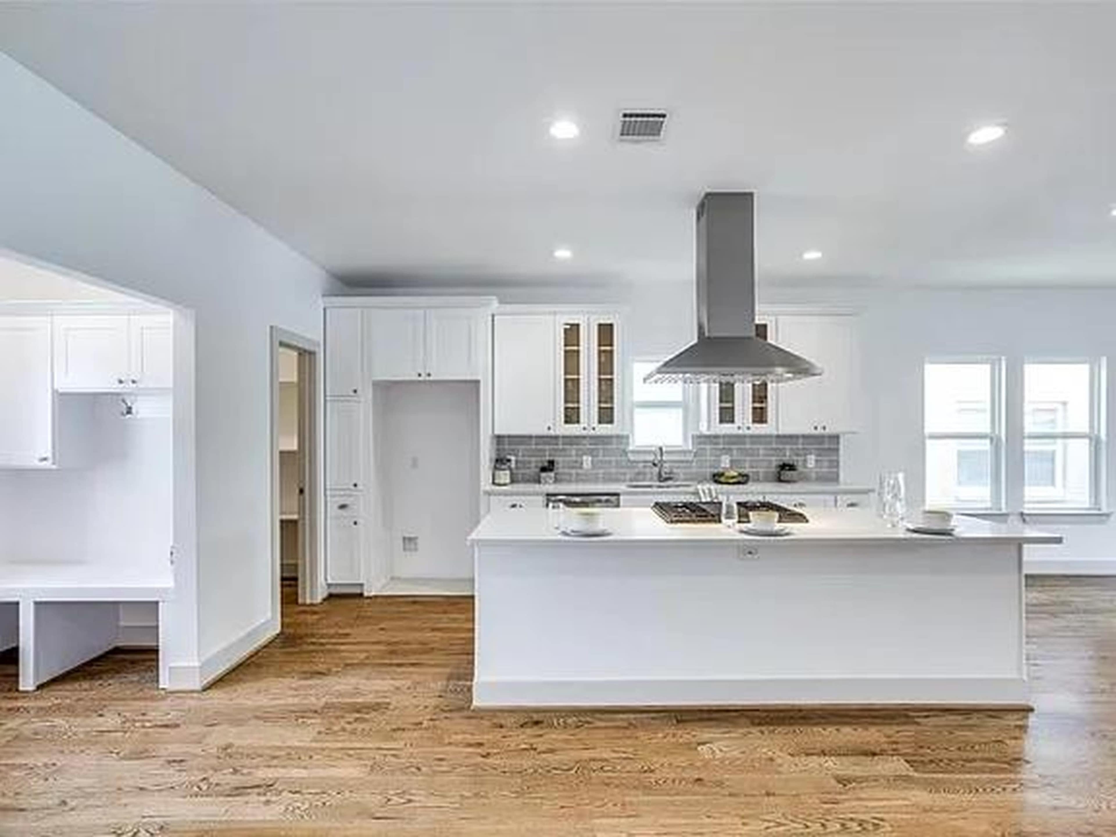 The image shows a modern kitchen with an island, stainless steel appliances, and white cabinetry, featuring hardwood floors and ample natural light.