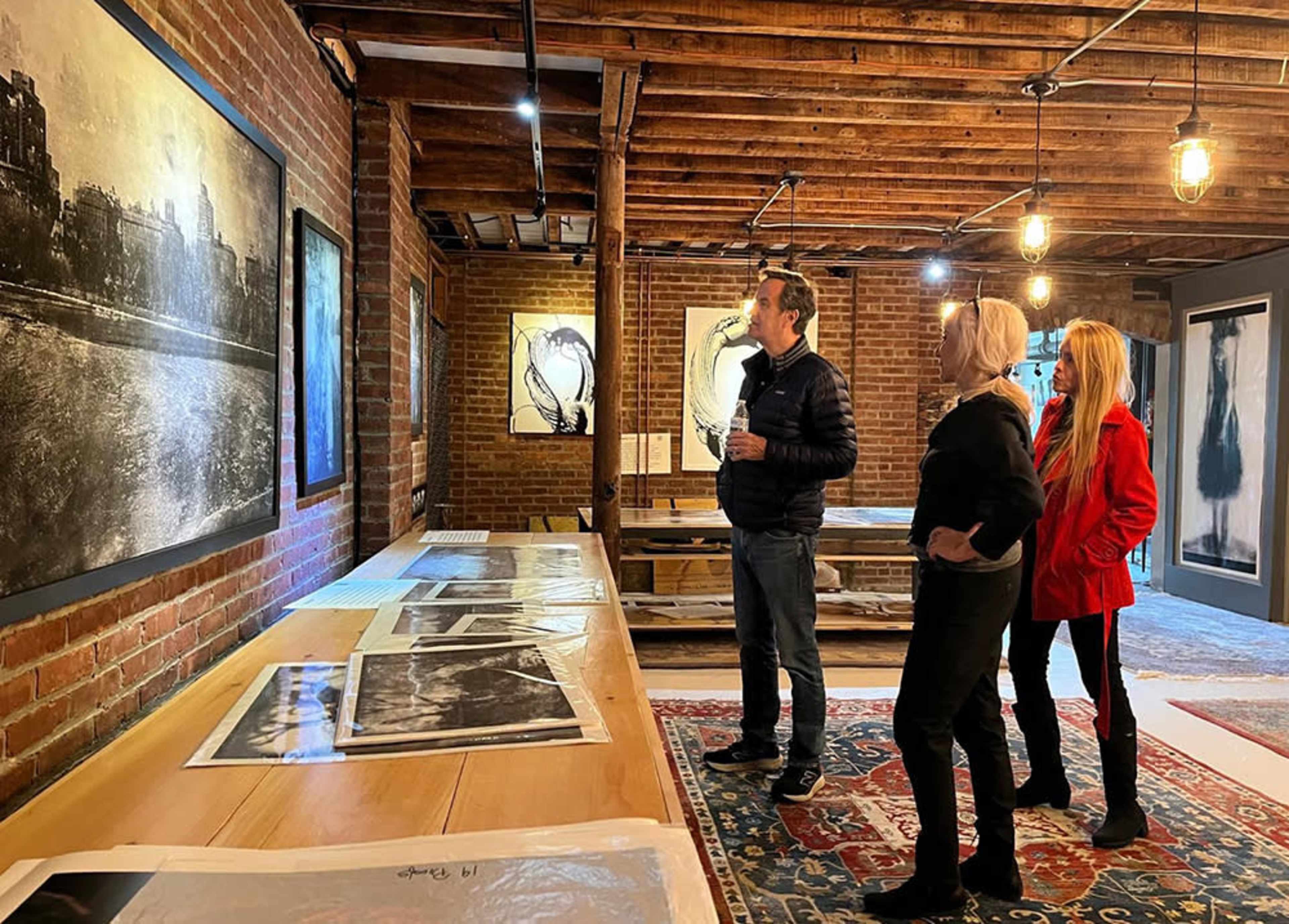 Three people examine artwork displayed in a gallery with exposed brick walls and wooden beams.