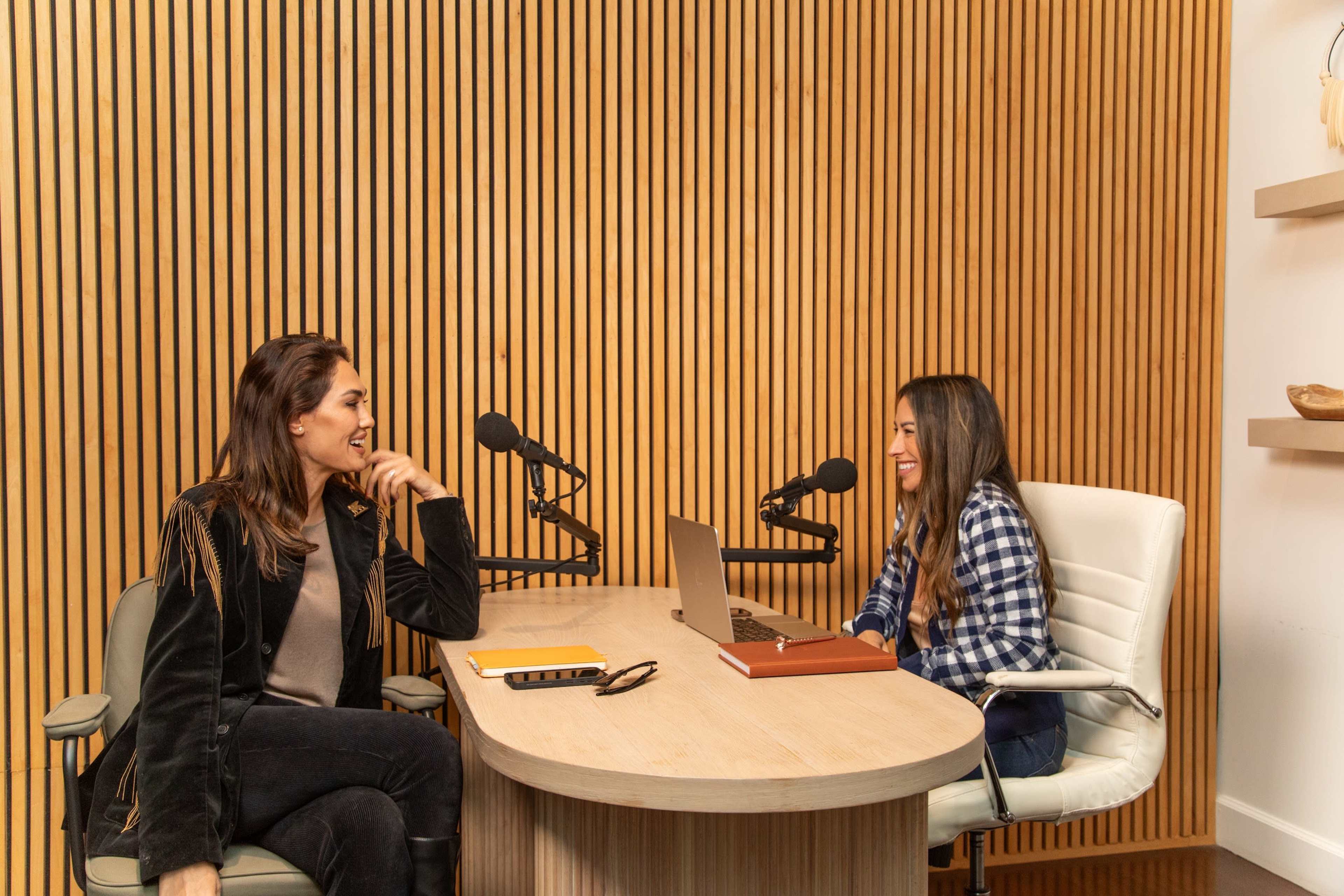 Two people are seated at a wooden table in a recording studio equipped with microphones, laptops, and notes.
