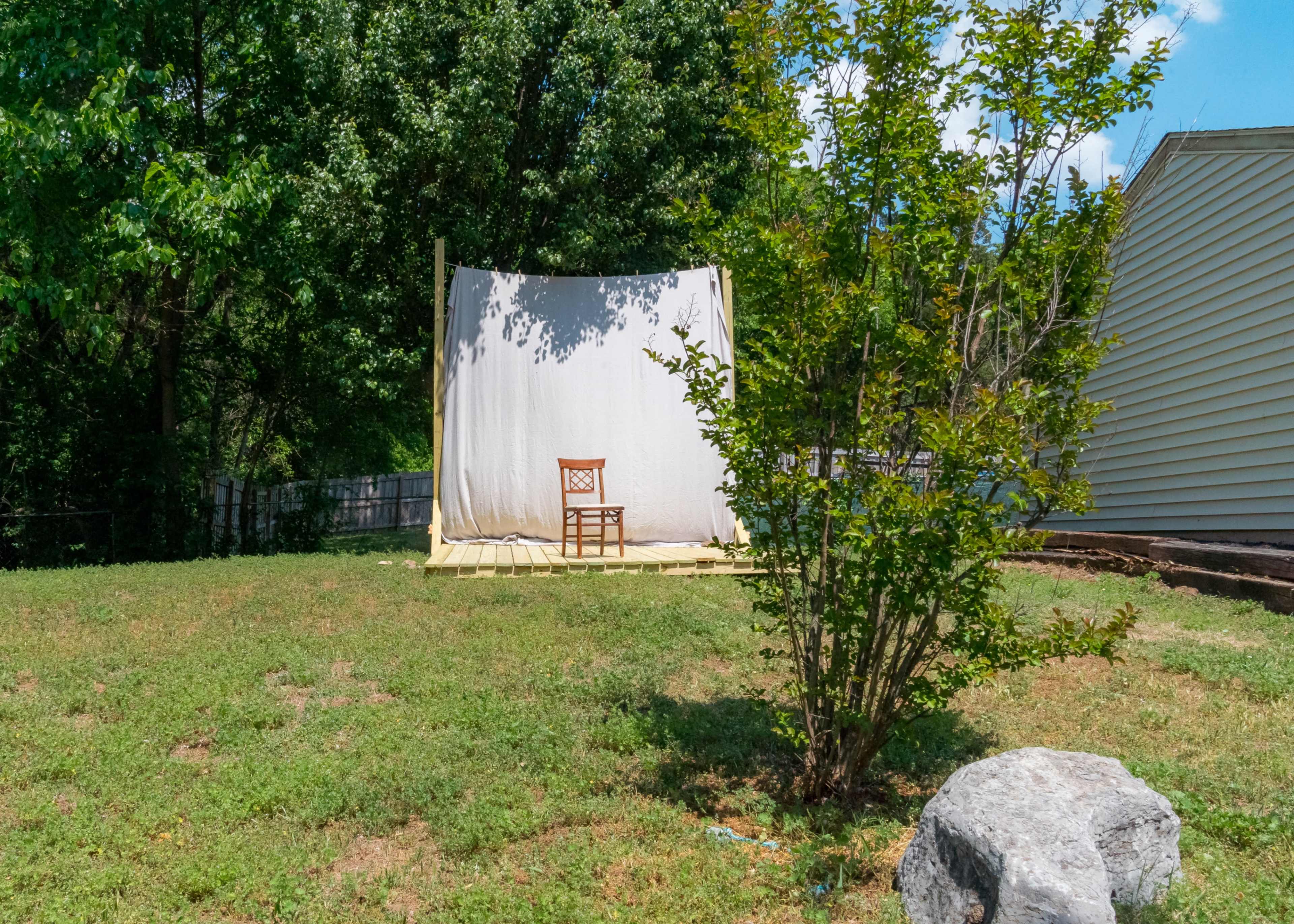 A wooden chair is positioned in front of a white fabric backdrop on a grassy area, surrounded by trees and a small bush.