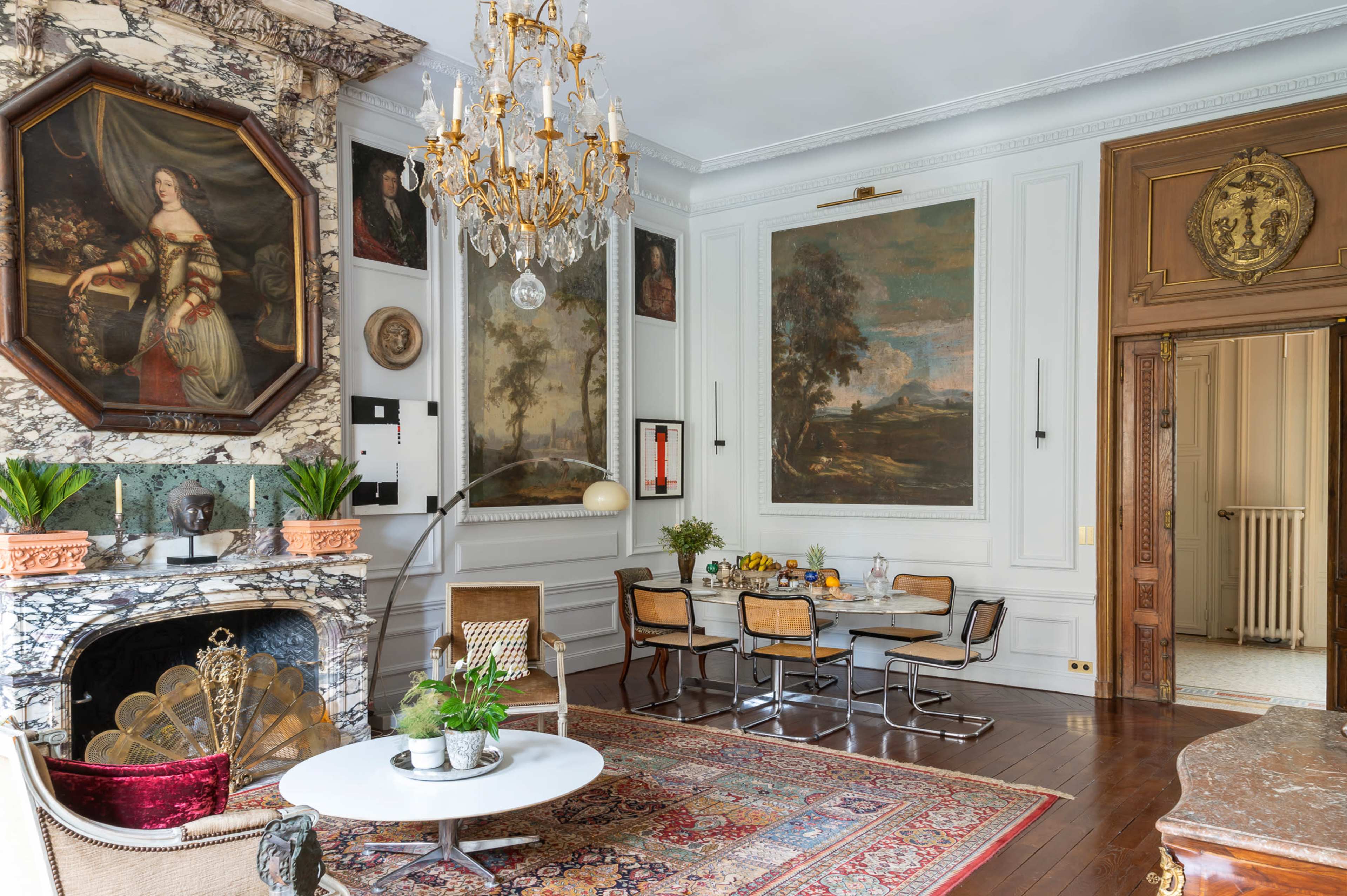 The room features a marble fireplace, a crystal chandelier, and a dining area with a table surrounded by modern chairs, all set against a backdrop of classical paintings and ornate moldings.