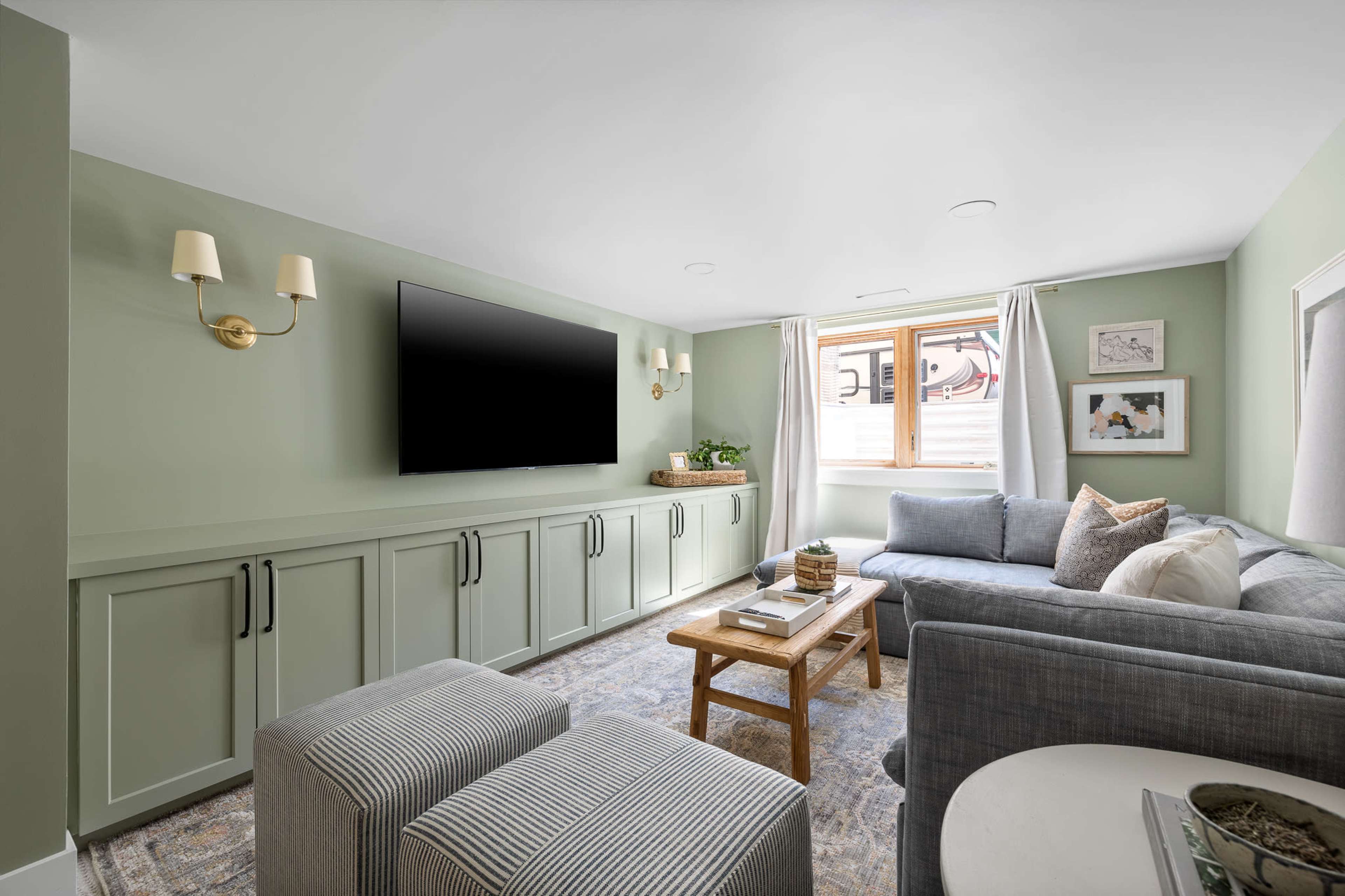 The living room features a sectional sofa, a coffee table, a wall-mounted television, and light green walls, with ample natural light from a window.