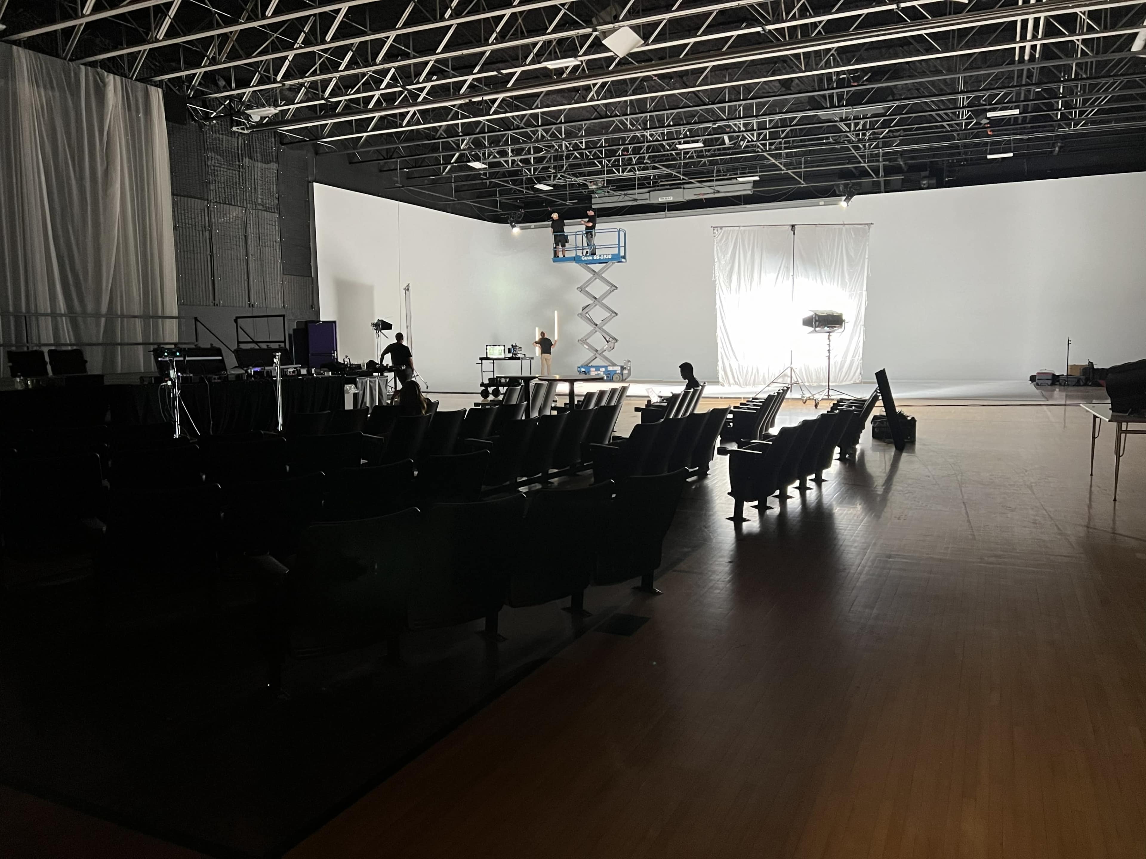 A large empty studio space features rows of black chairs facing a well-lit backdrop, with filming equipment positioned throughout.