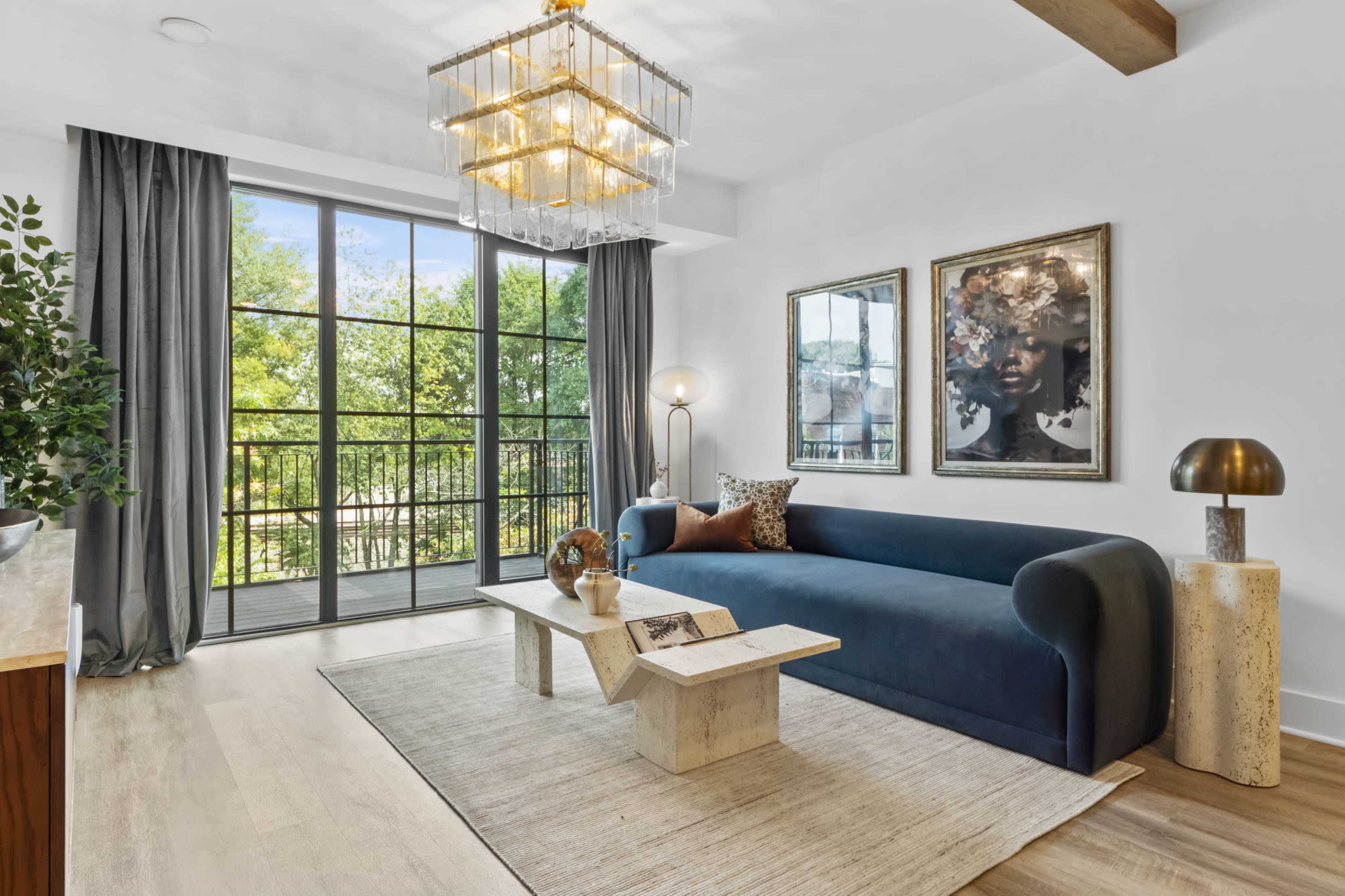 The image shows a modern living room featuring a blue sofa, a coffee table made of stone, and large windows with views of greenery outside.