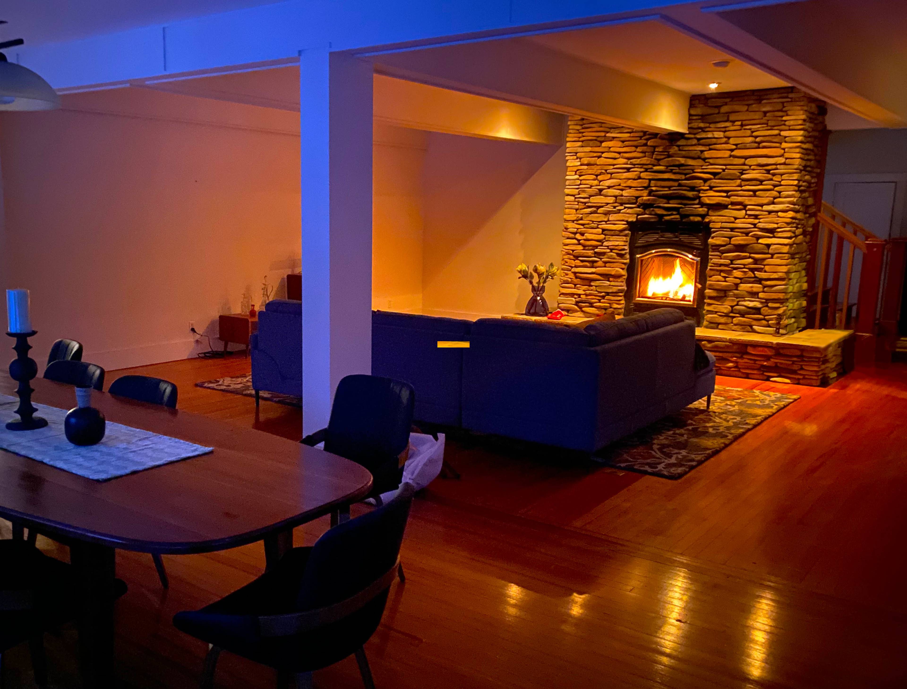 A living room features a stone fireplace surrounded by a sofa and wooden dining table, illuminated by soft lighting.