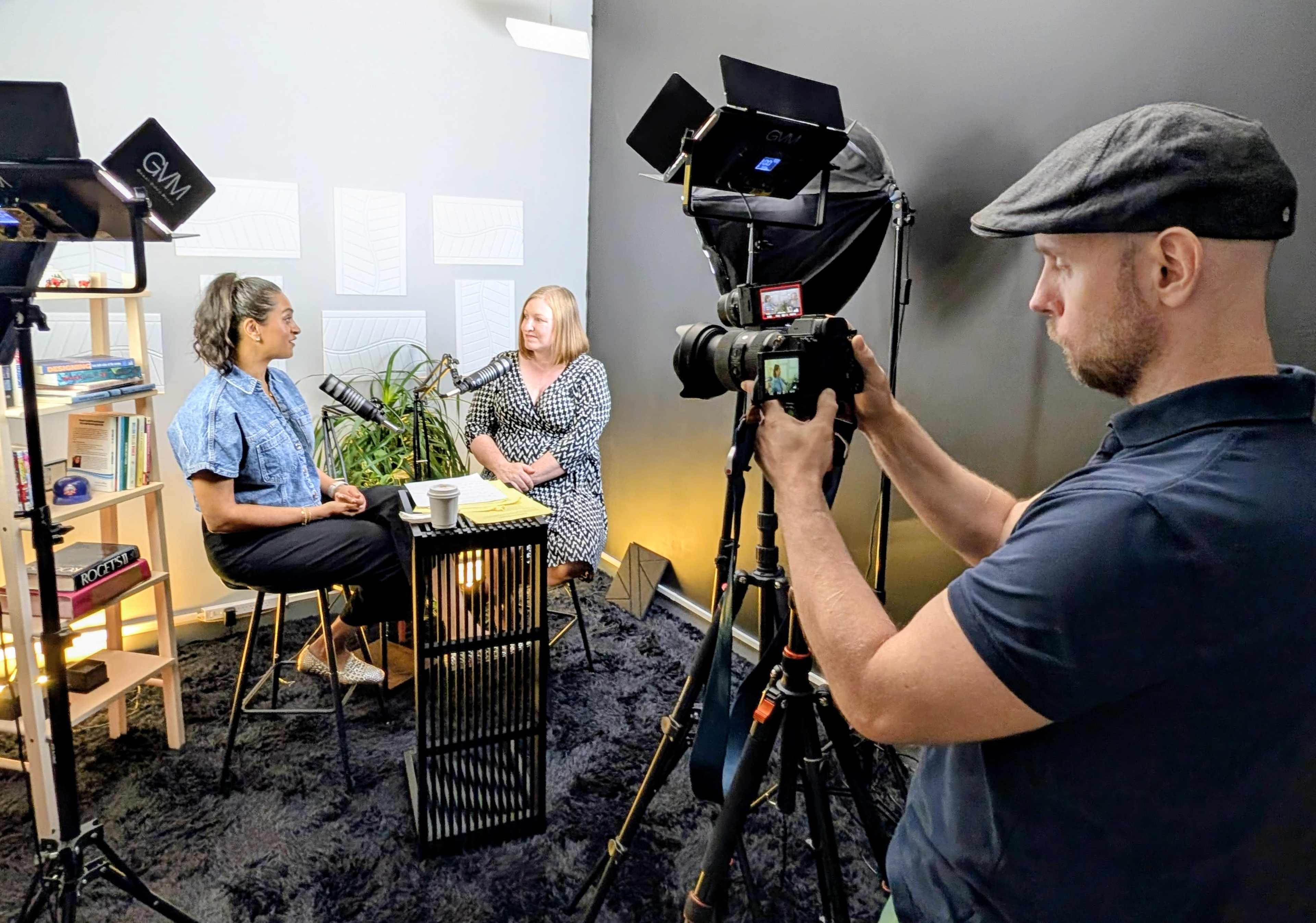 A camera operator is filming an interview between two women seated at a table in a well-lit, stylish studio setting.