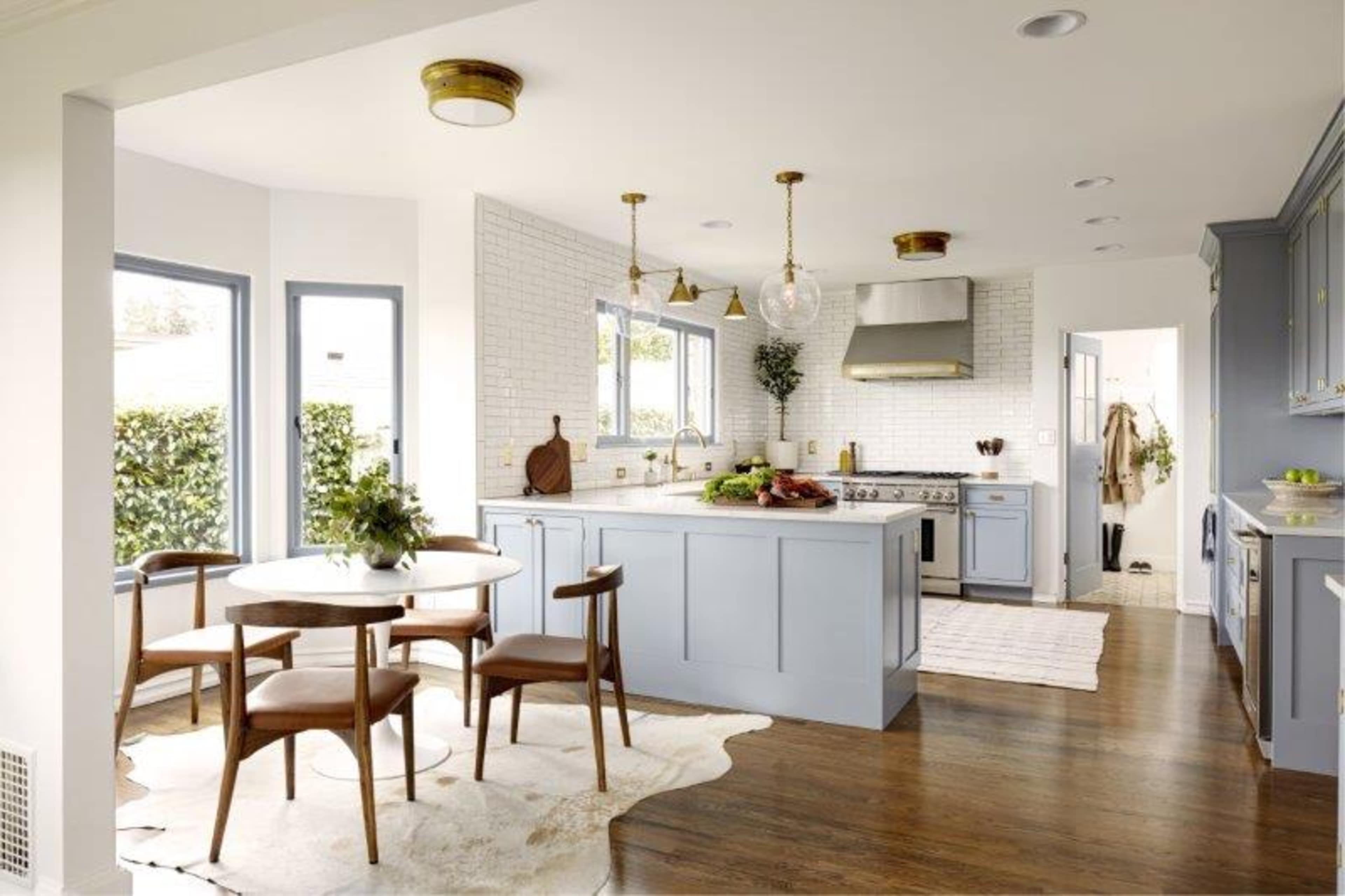 A modern kitchen features light blue cabinetry, a wooden dining table with four chairs, and large windows allowing natural light.