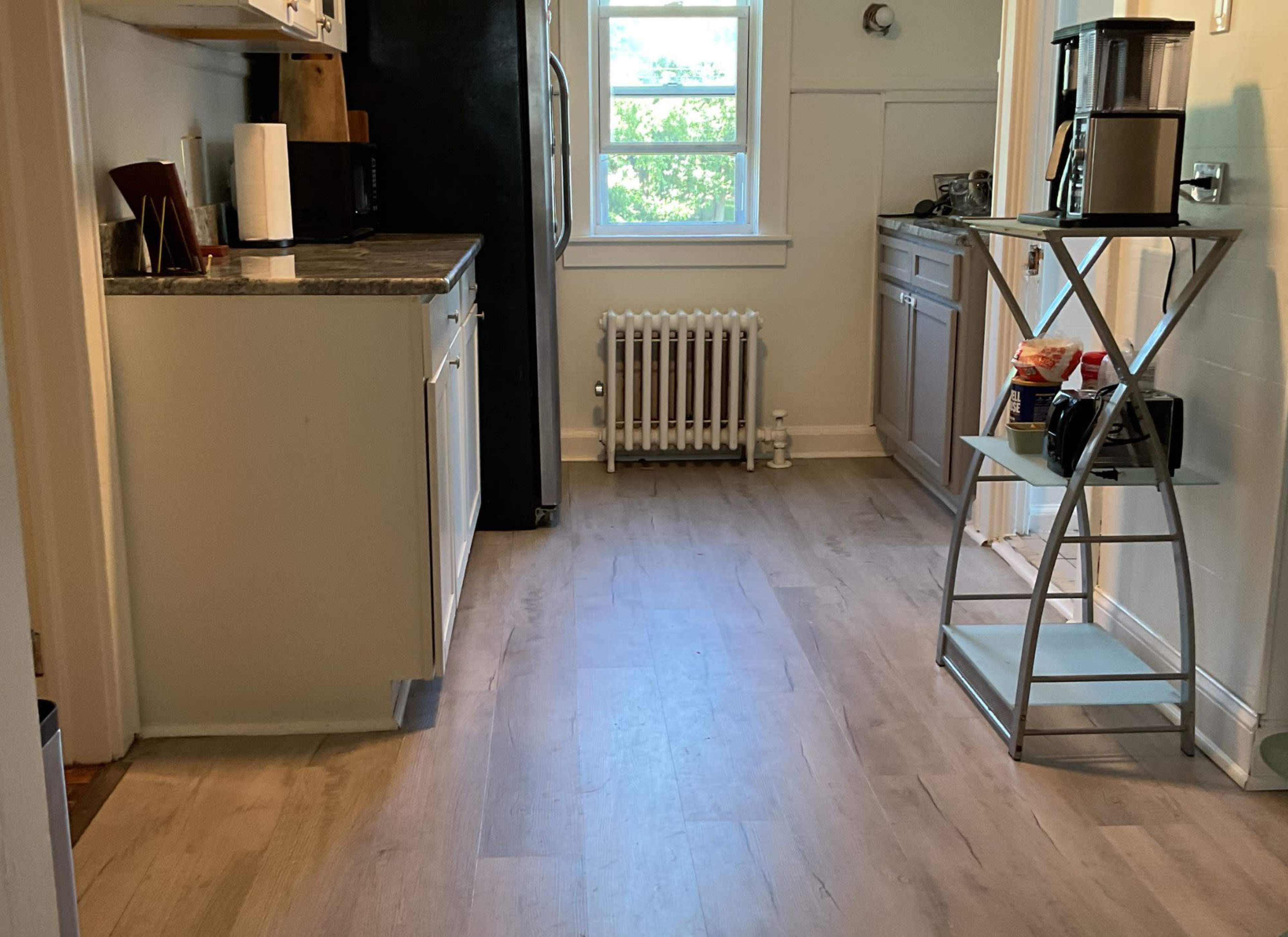 The image shows a kitchen with a light-colored floor, a black refrigerator, white cabinets, and a metal cart holding a coffee maker and snacks.