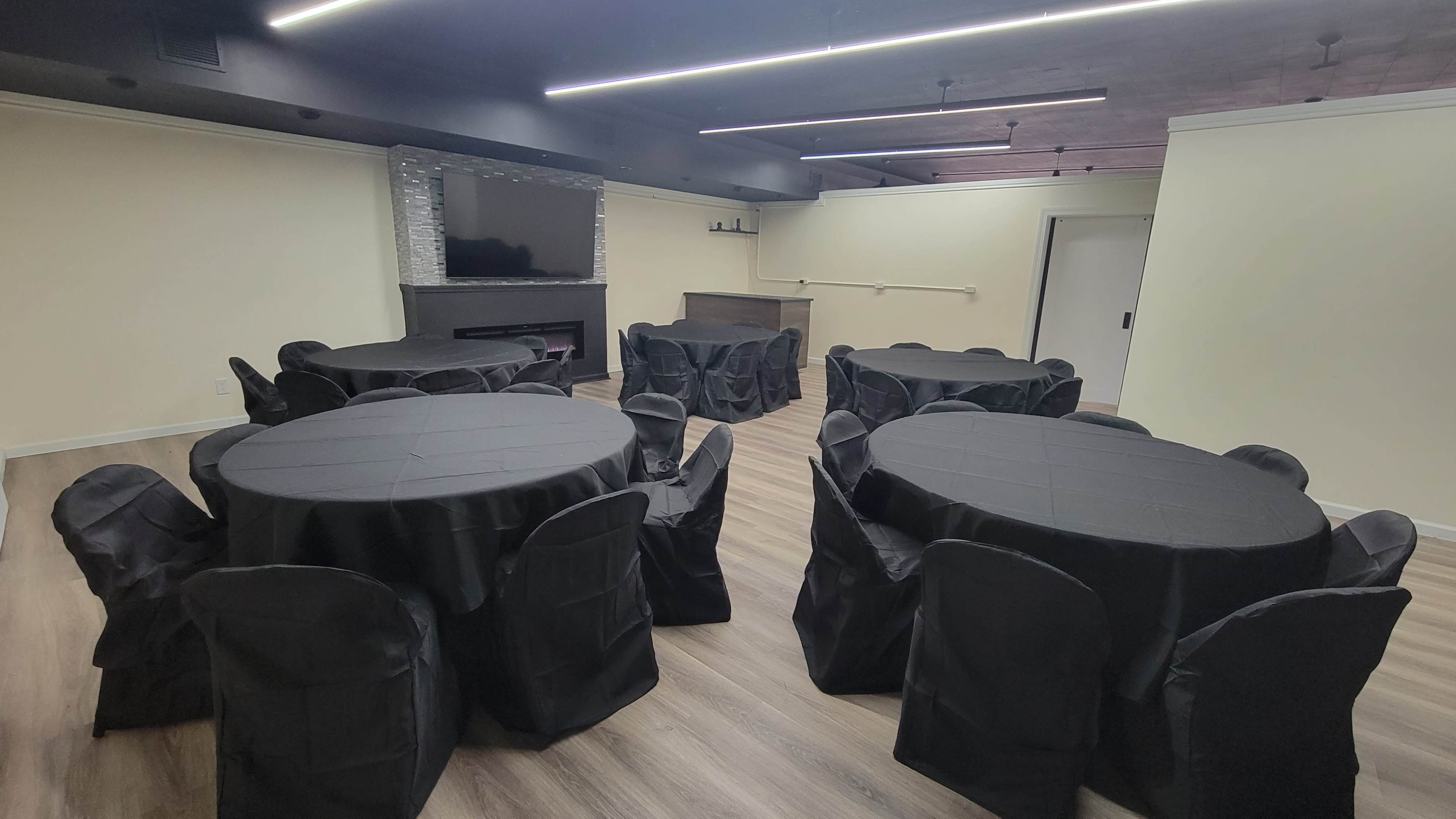 The image shows a room set up for an event, featuring several round tables covered with black tablecloths and matching chairs arranged around them.