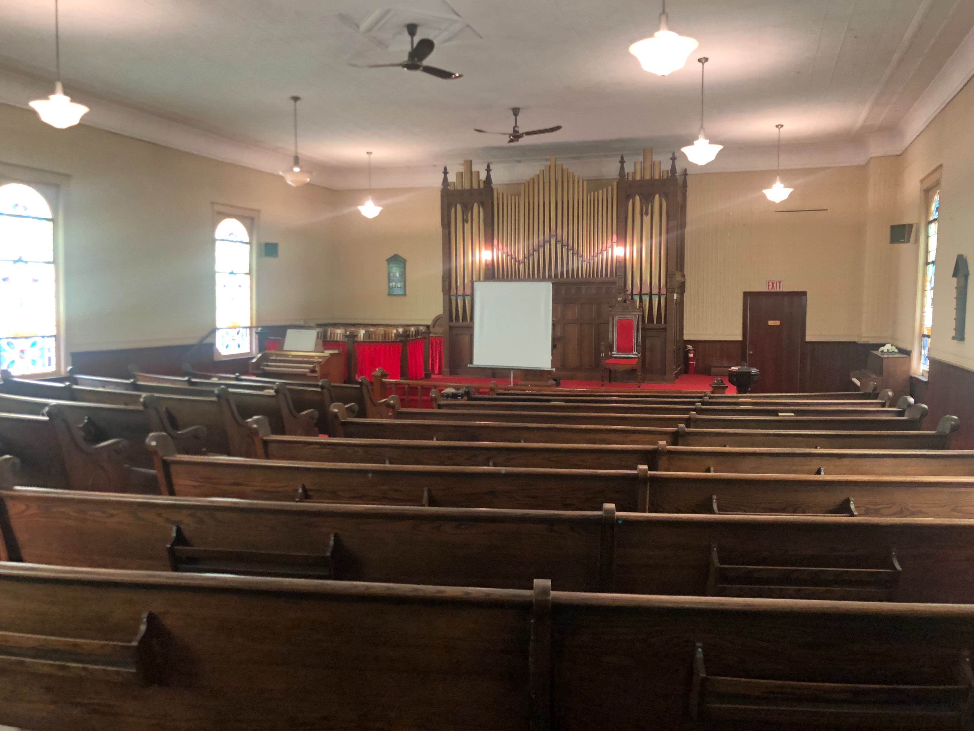 The interior of a church features wooden pews facing a stage with an organ, red furnishings, and a projector screen set up for a presentation.