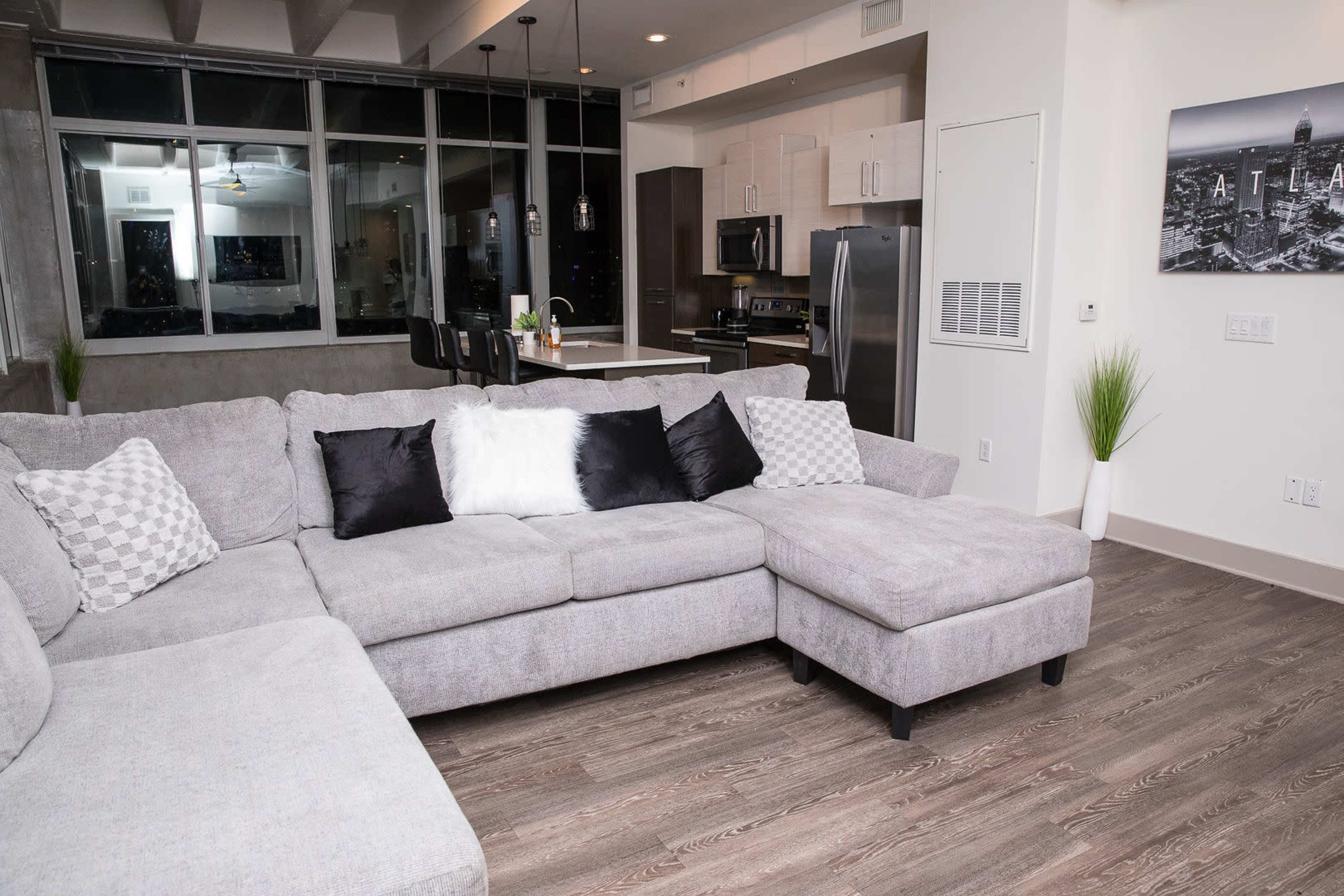 A modern living room features a gray sectional sofa, a small ottoman, and a kitchen in the background with black appliances and bar seating.