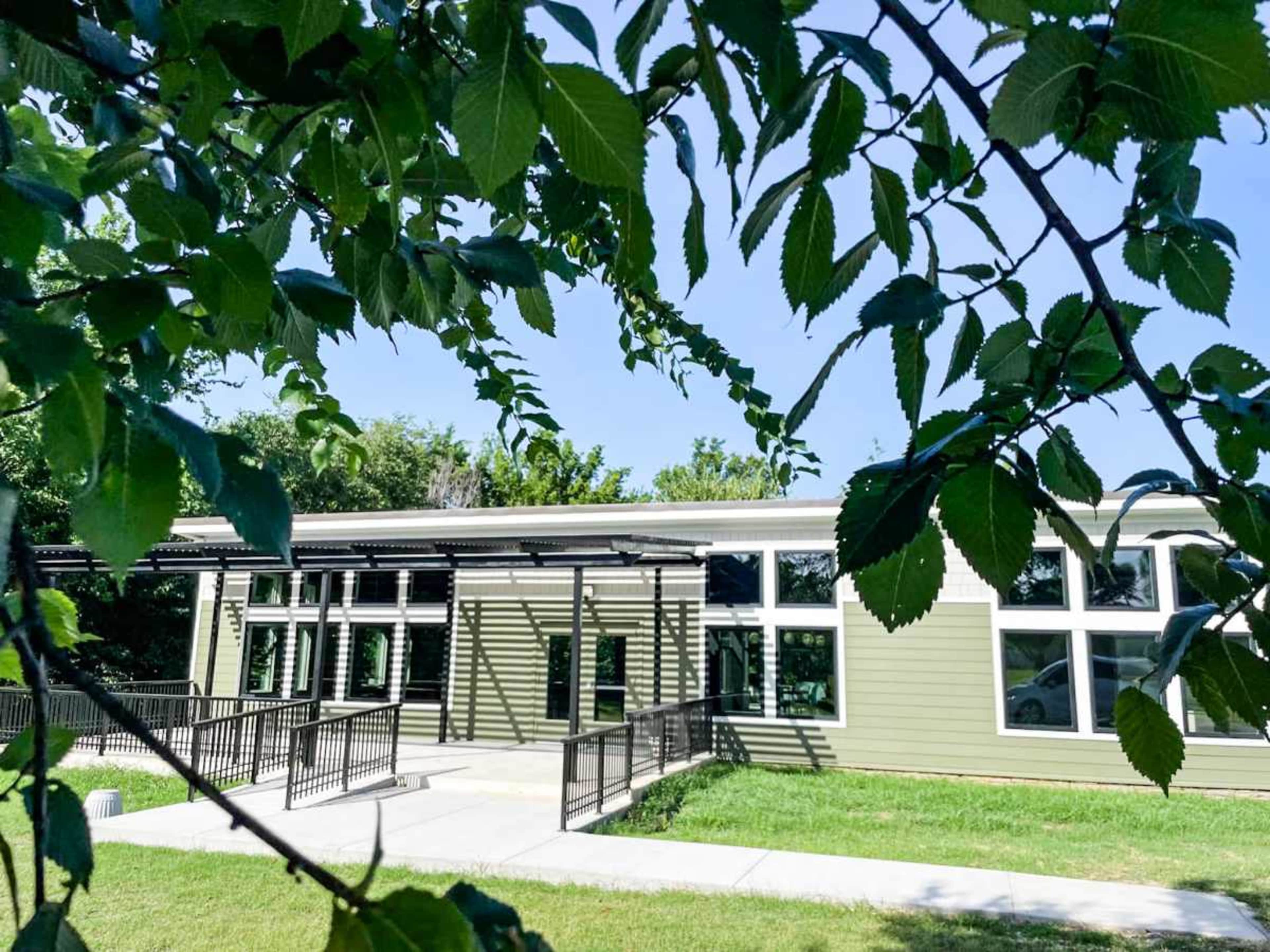 A green building with large windows and a ramp is partially obscured by leafy branches in a sunny outdoor setting.