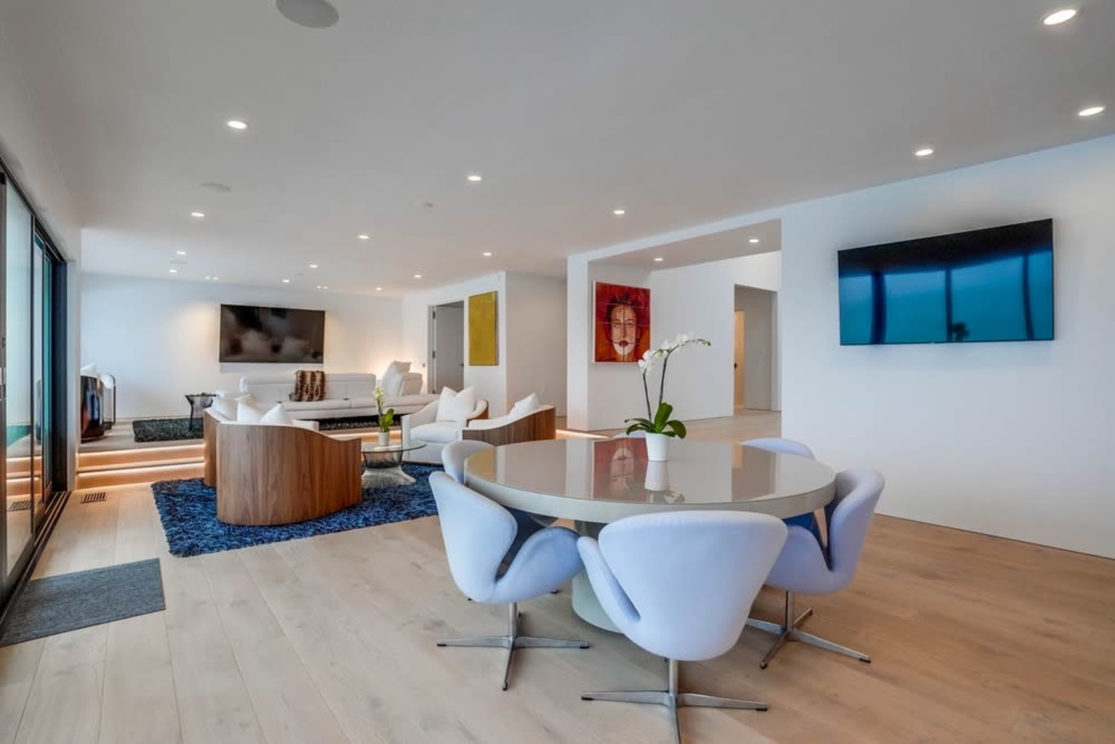 The image shows a modern living space featuring a dining area with a round table surrounded by upholstered chairs, a seating area with a sofa and television, and large windows allowing natural light.