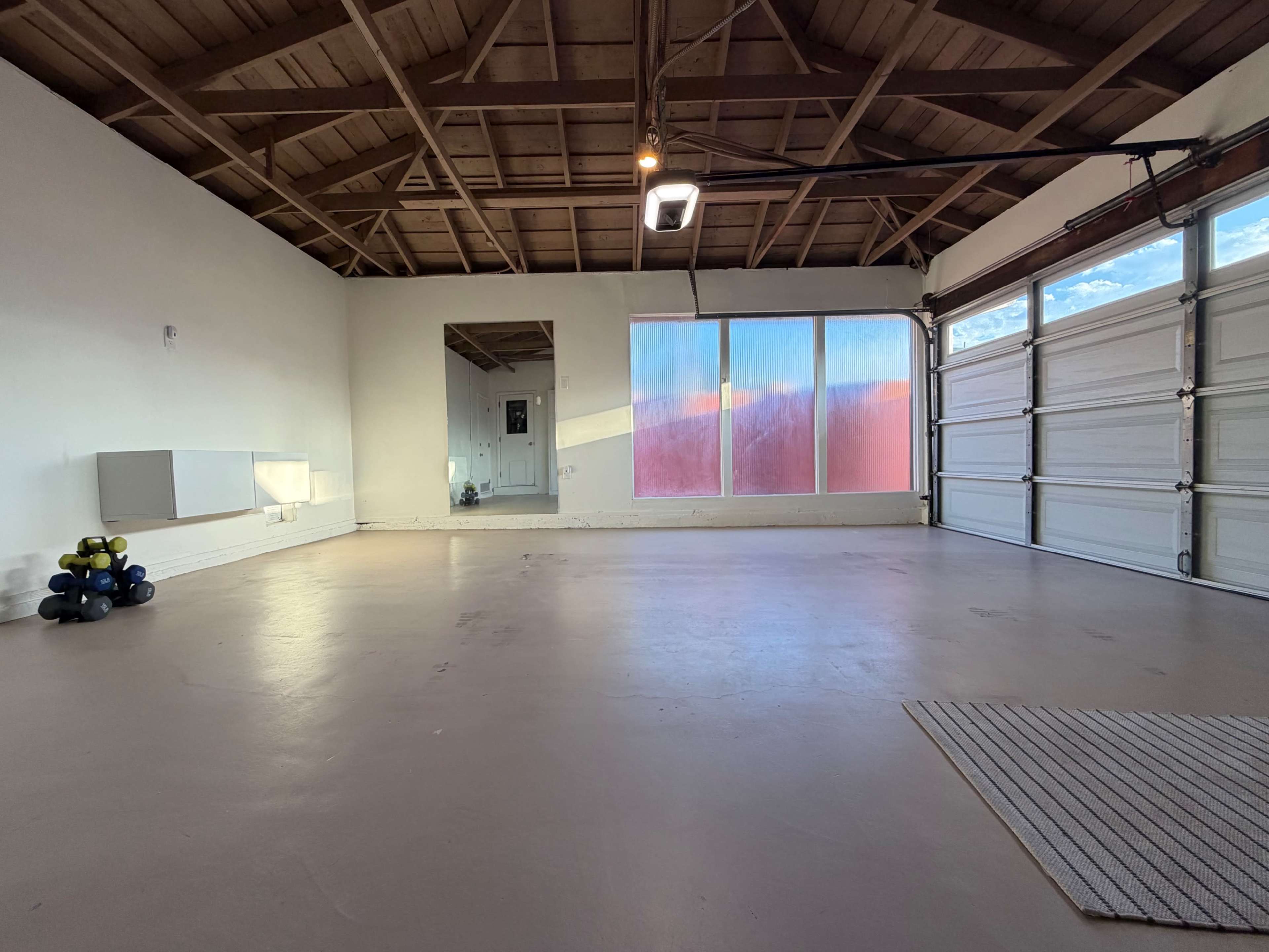 The image shows an empty garage with a wooden ceiling, a large window, and a few exercise weights in the corner.