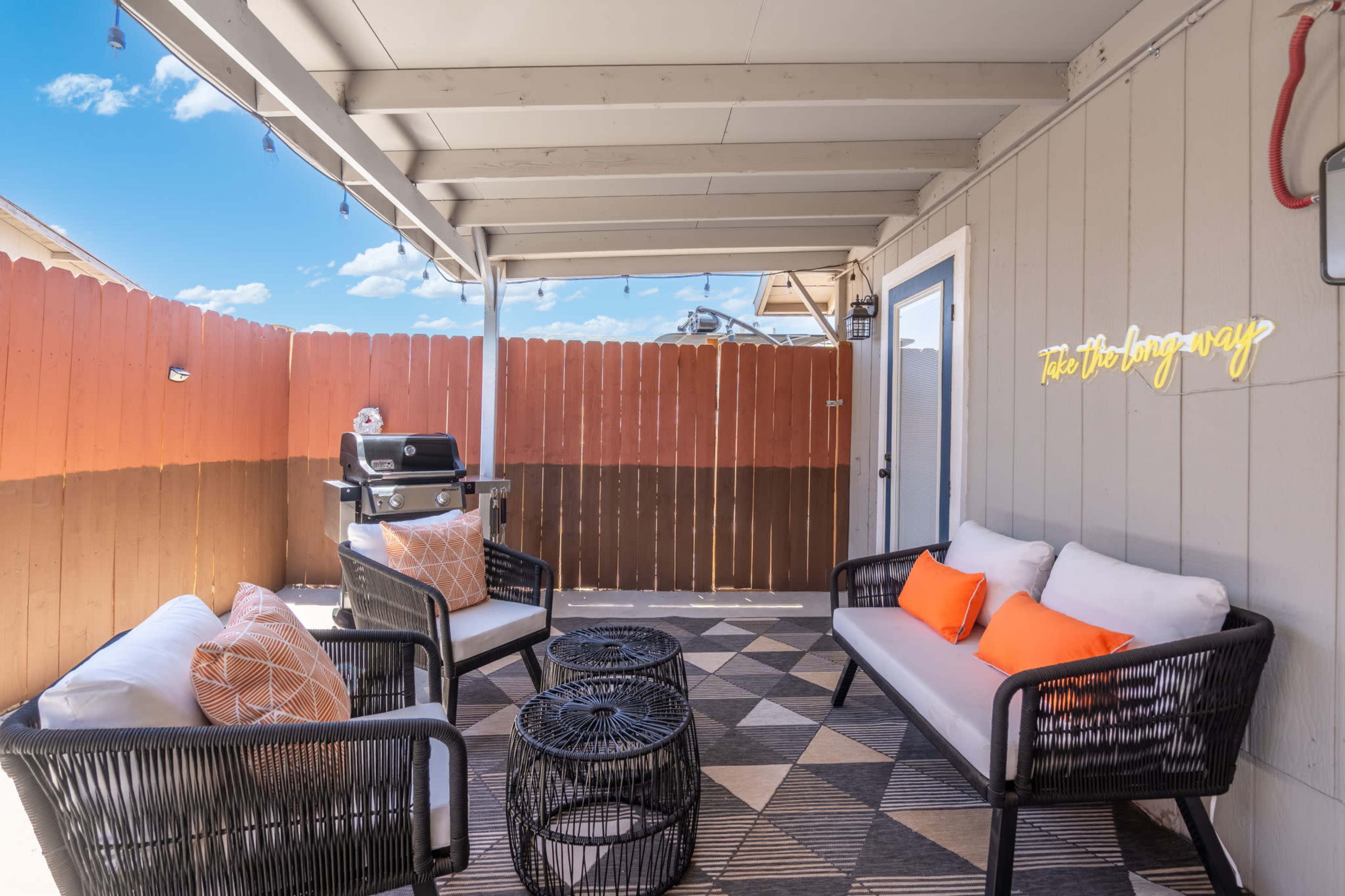 The image shows a cozy outdoor patio area with two seating arrangements, a small table, and a barbecue grill, all enclosed by a wooden fence.