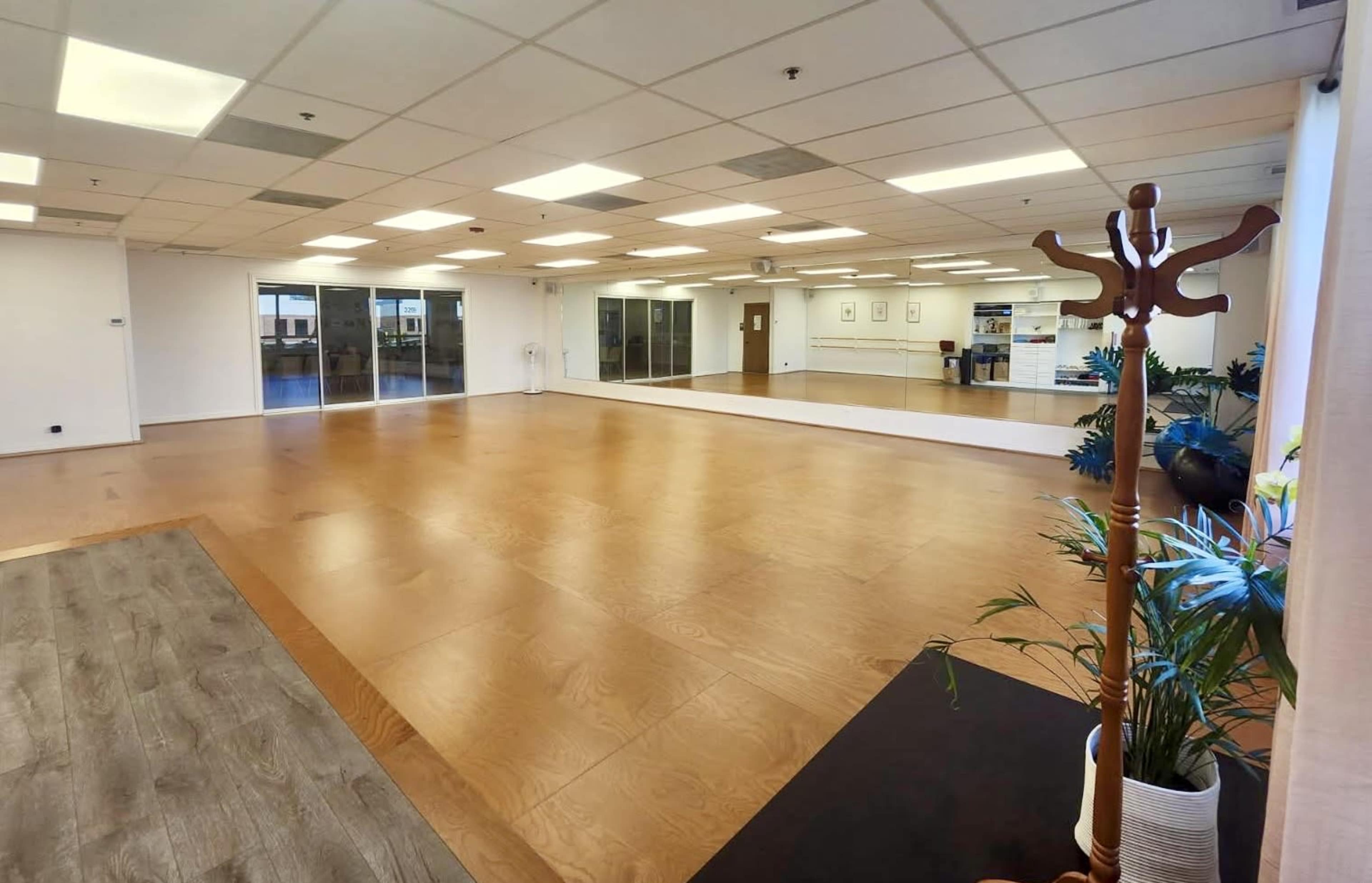 The image shows a spacious, well-lit studio with wooden flooring, large windows, and a mirrored wall.