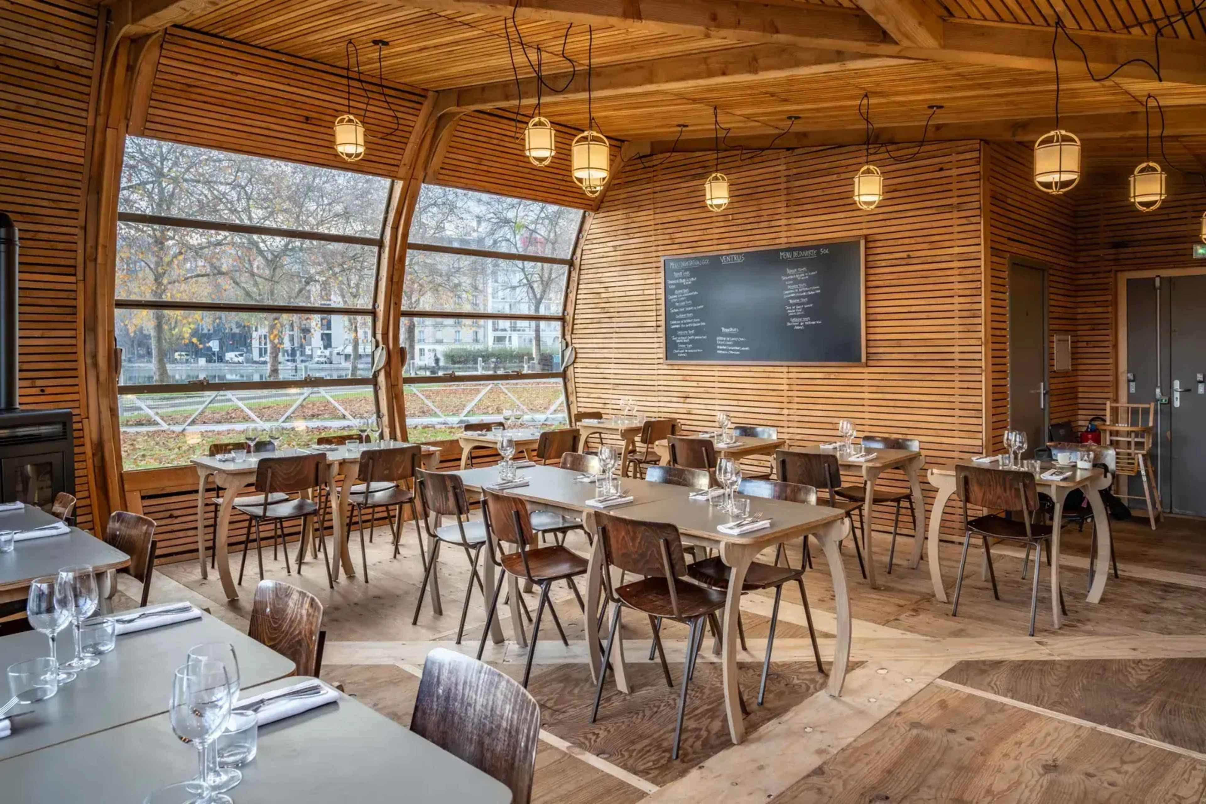 A cozy restaurant interior featuring wooden walls, tables set for dining, and large windows overlooking an outdoor view.