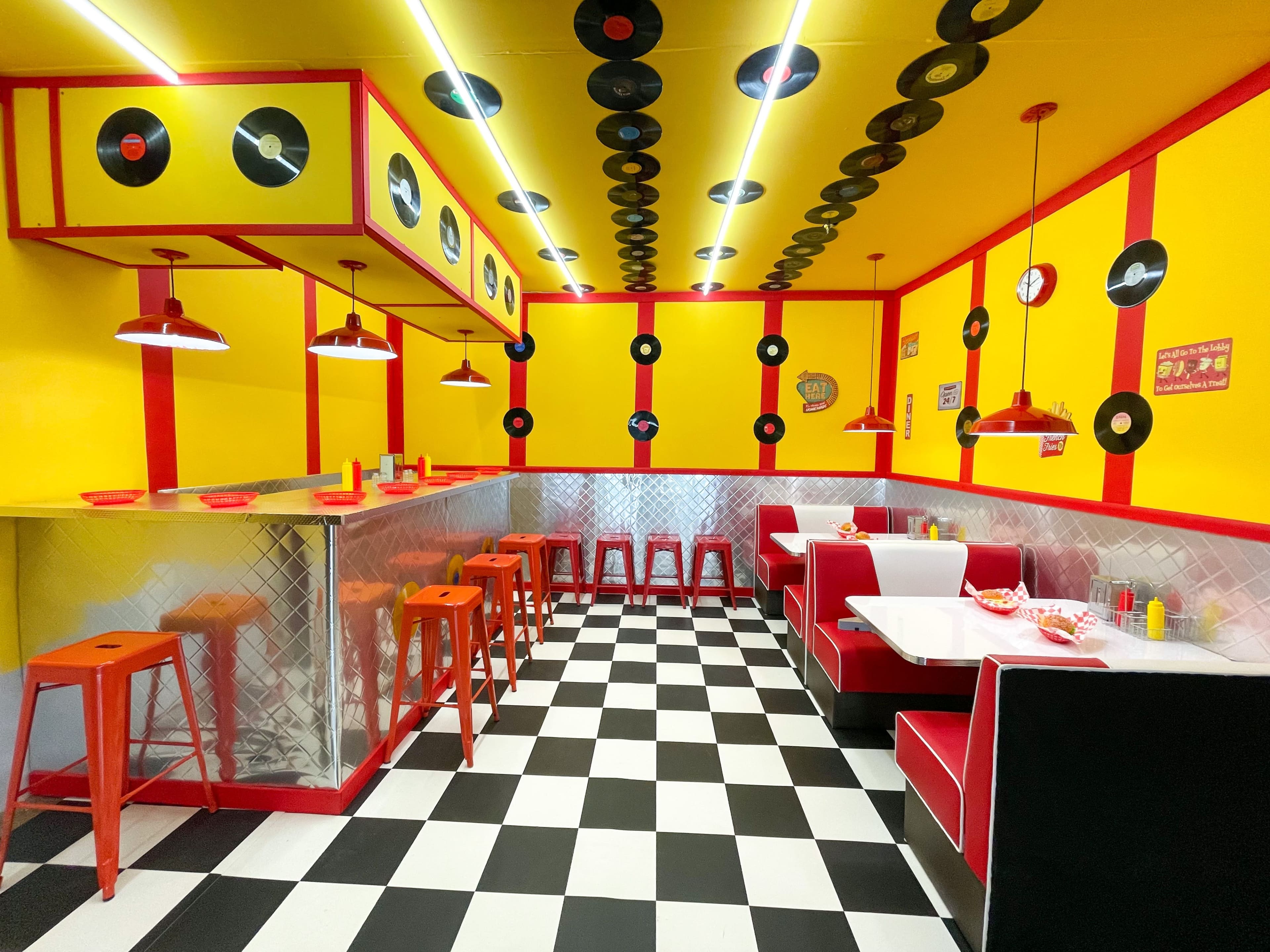 A vintage-style diner features yellow walls adorned with records, red accents, a checkered floor, and both bar and booth seating.