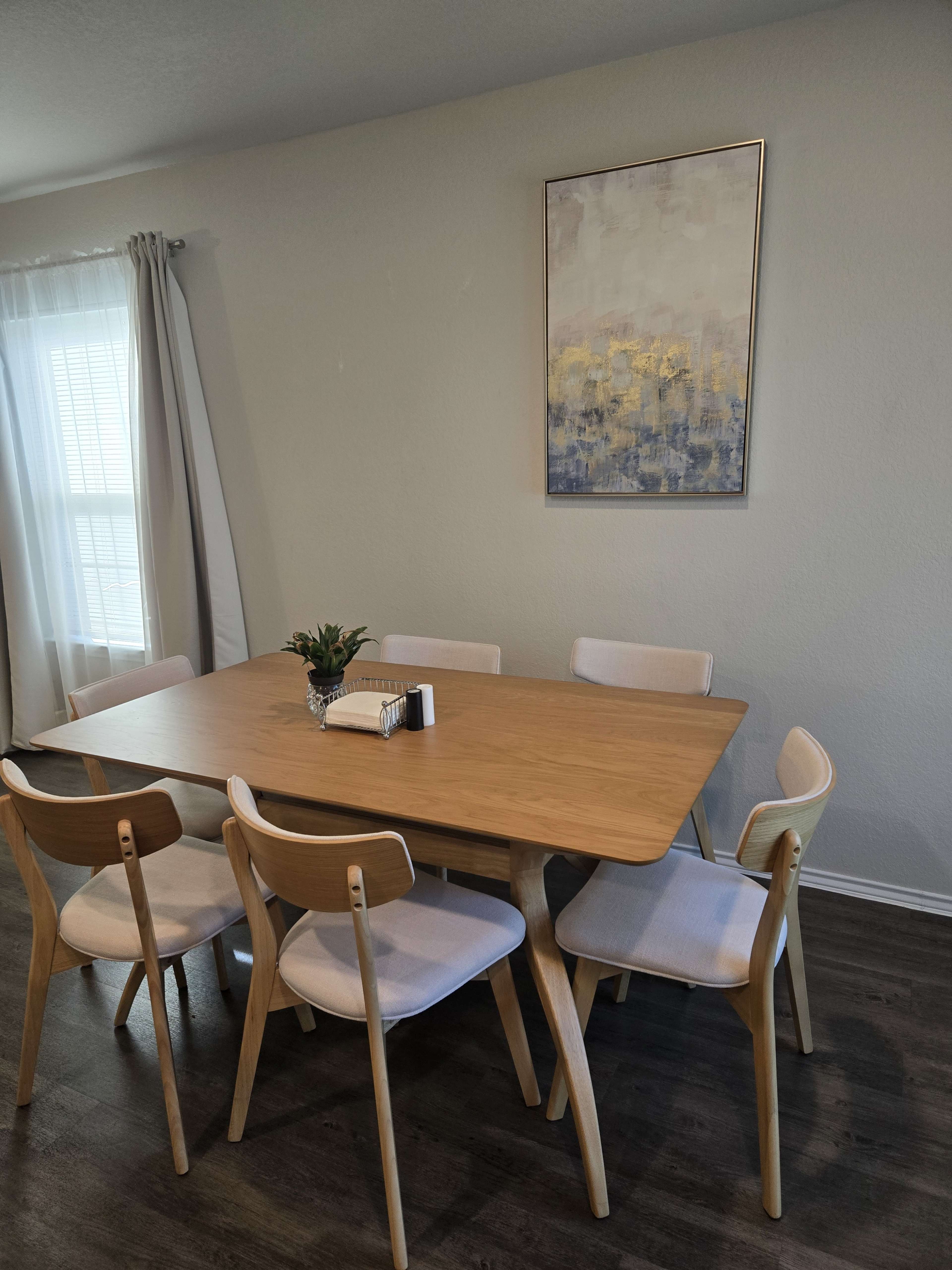 The image shows a wooden dining table with six chairs arranged around it, situated in a room with light-colored walls and a piece of abstract art hanging on the wall above the table.