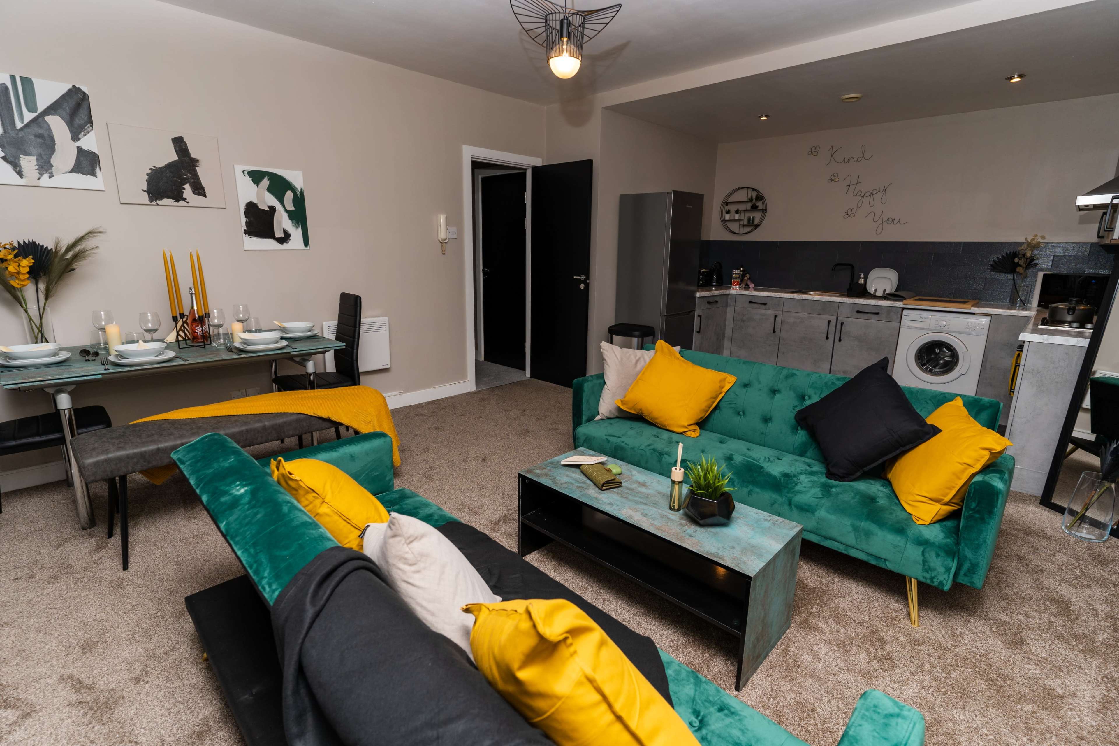 A modern living area featuring a seating arrangement with green and black sofas, a dining table set for four, and a kitchen area in the background.