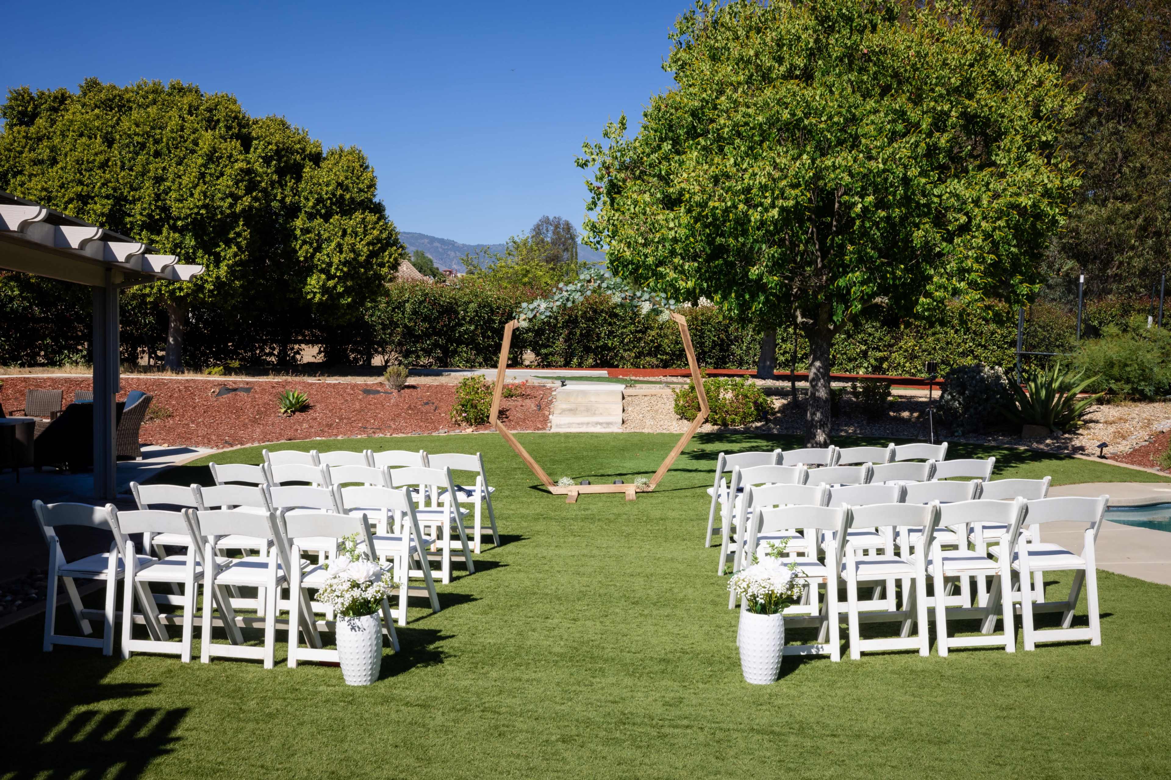 A wedding ceremony setup features white chairs arranged in rows on green grass, with a wooden arch framed by trees in the background.