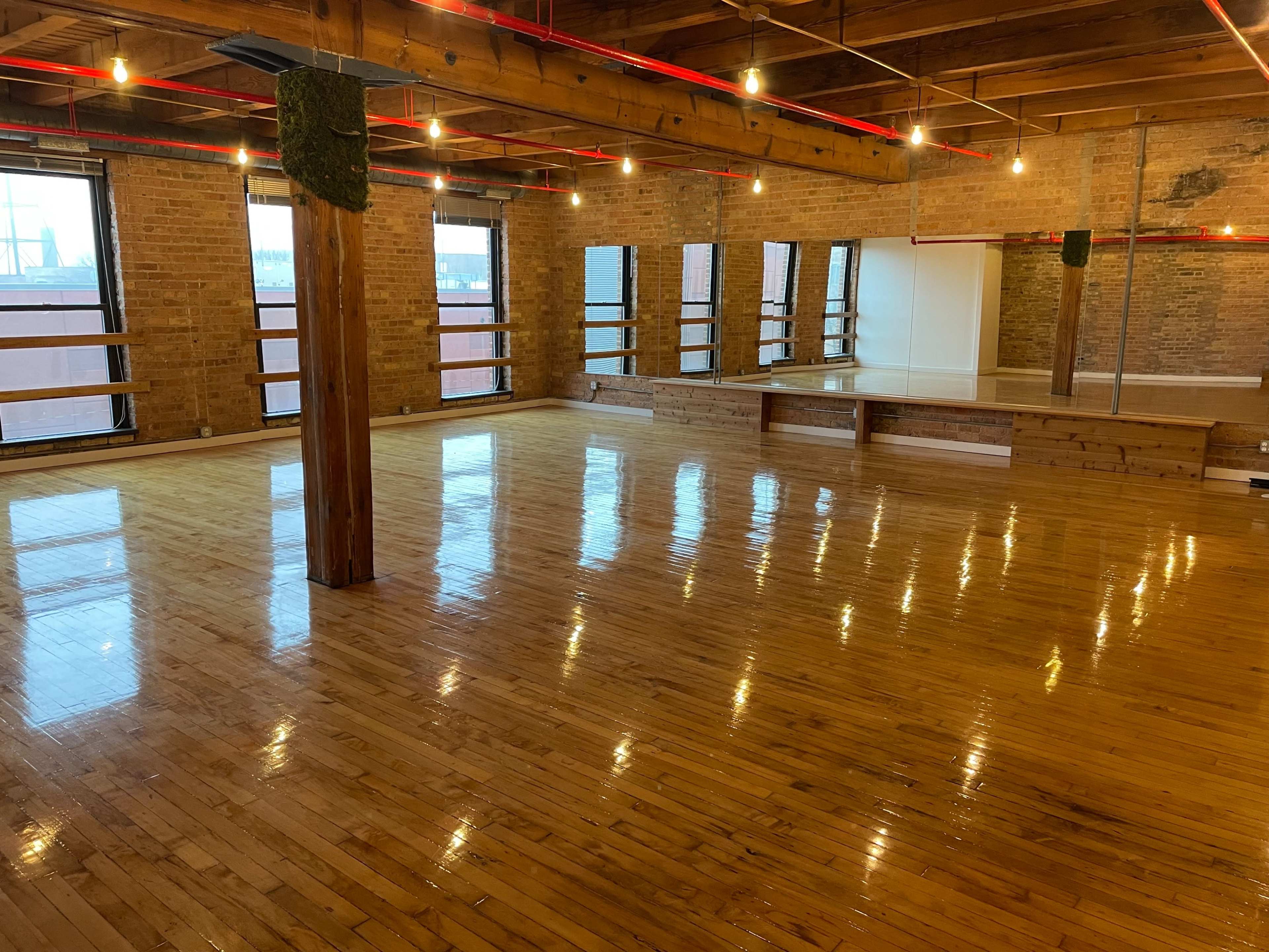 The image shows a spacious room with polished wooden floors, exposed brick walls, and large windows allowing natural light to enter.