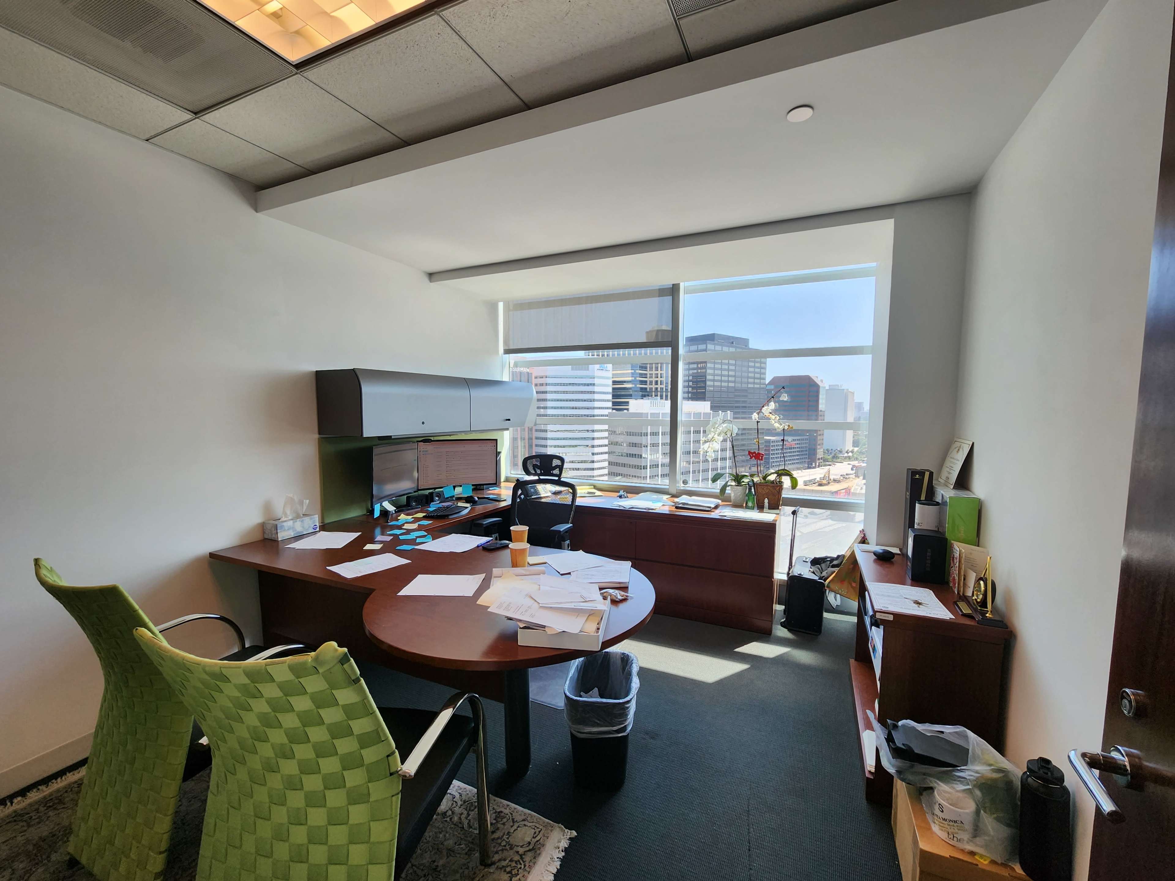 An office space features a wooden desk cluttered with papers, two green chairs, and a large window displaying a cityscape.
