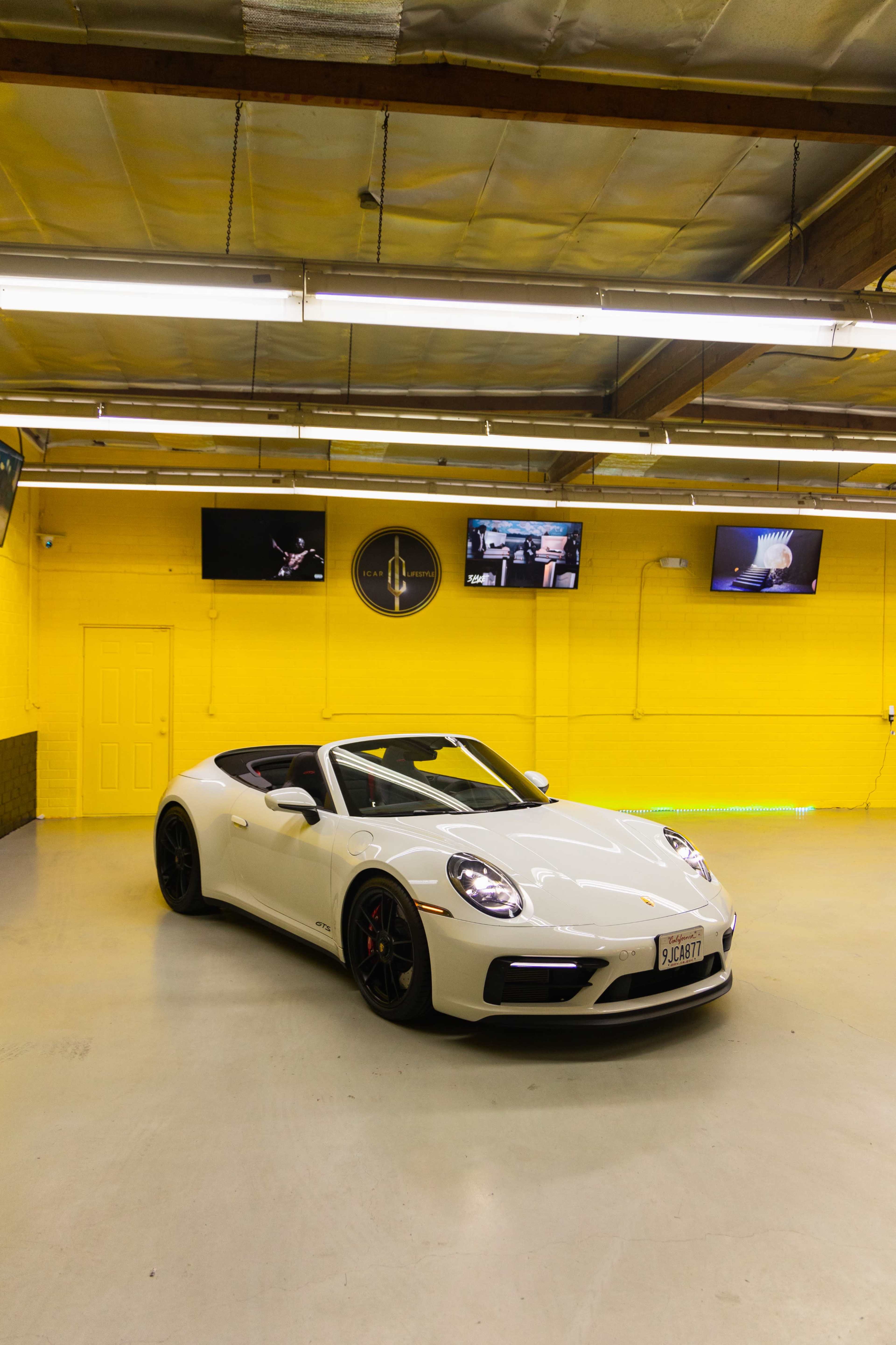 A white sports car is parked in a bright yellow garage with multiple screens mounted on the walls.
