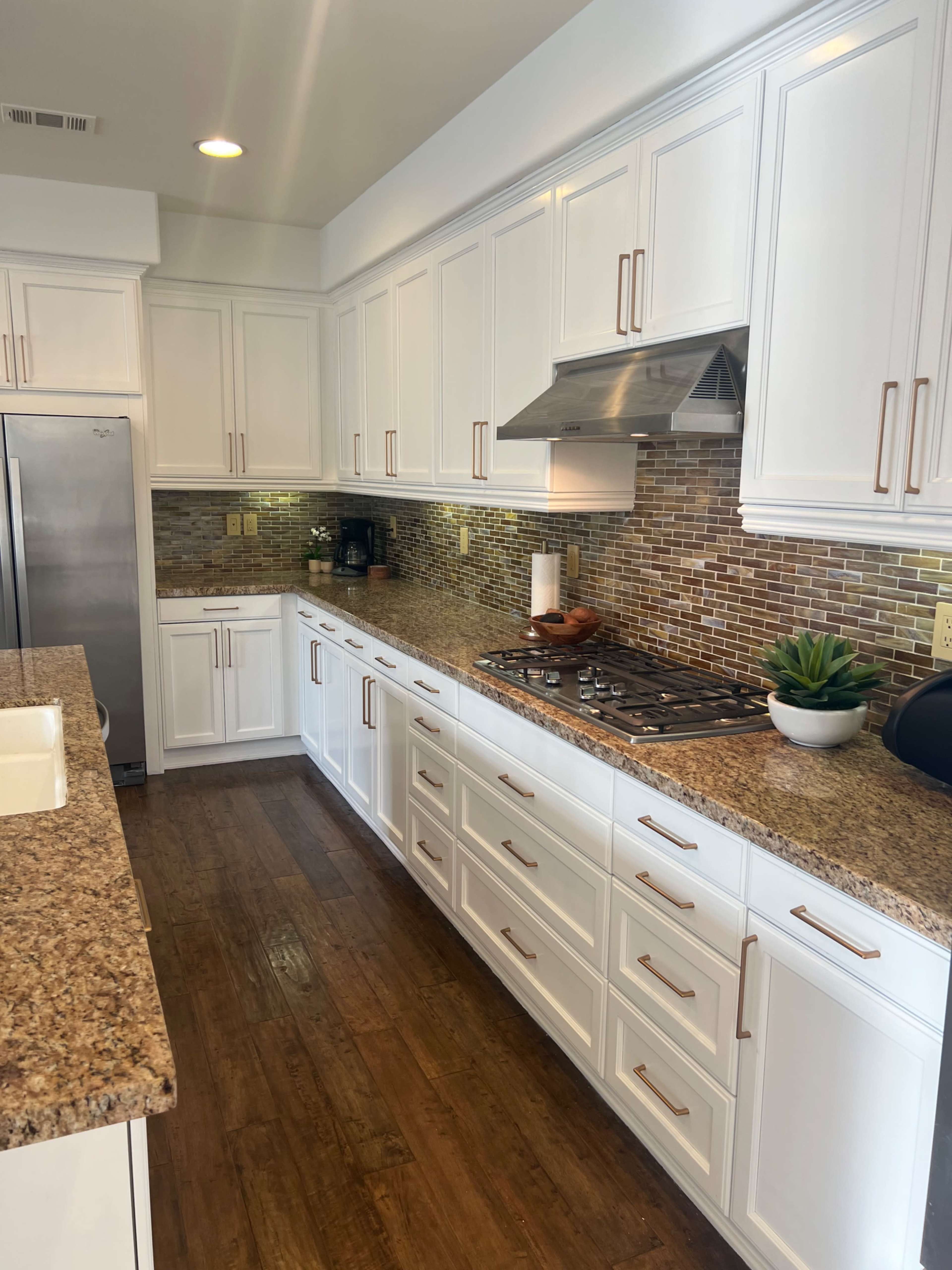 The image shows a modern kitchen with white cabinetry, a granite countertop, and a tile backsplash featuring brown and gold tones.
