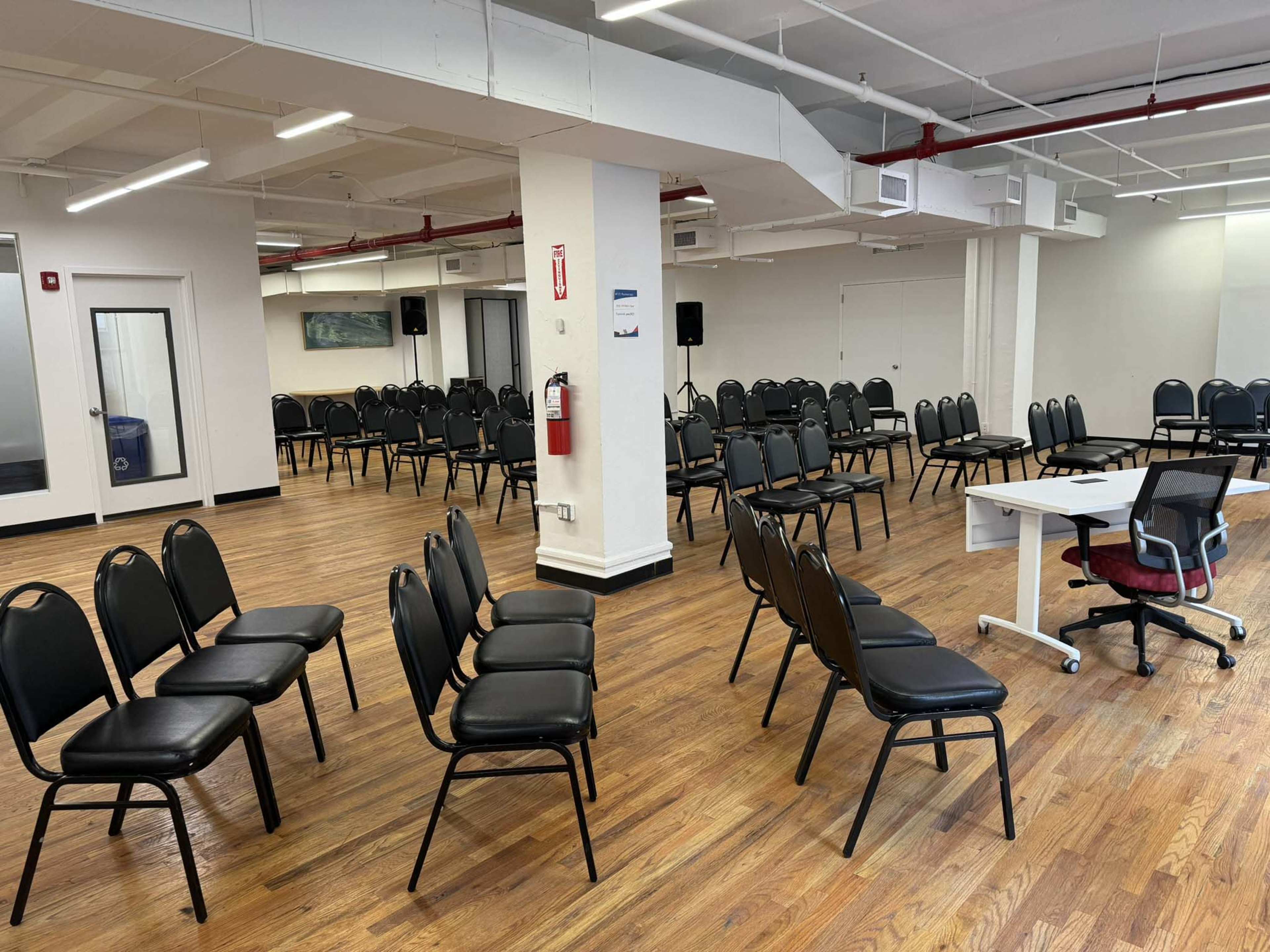 The image shows a spacious room with several rows of black chairs arranged in a semi-circle, and a single white table with a chair at one end.