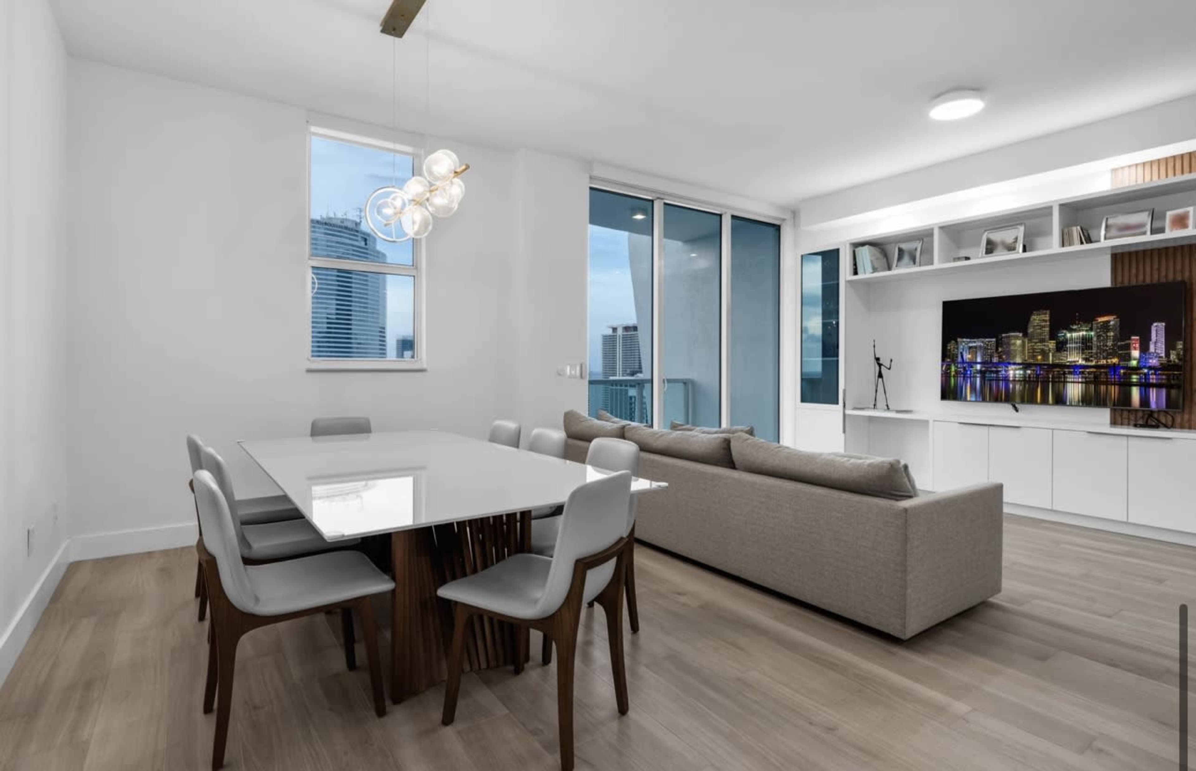 A modern dining area features a white table with six chairs, a gray sofa, and large windows overlooking a city skyline.