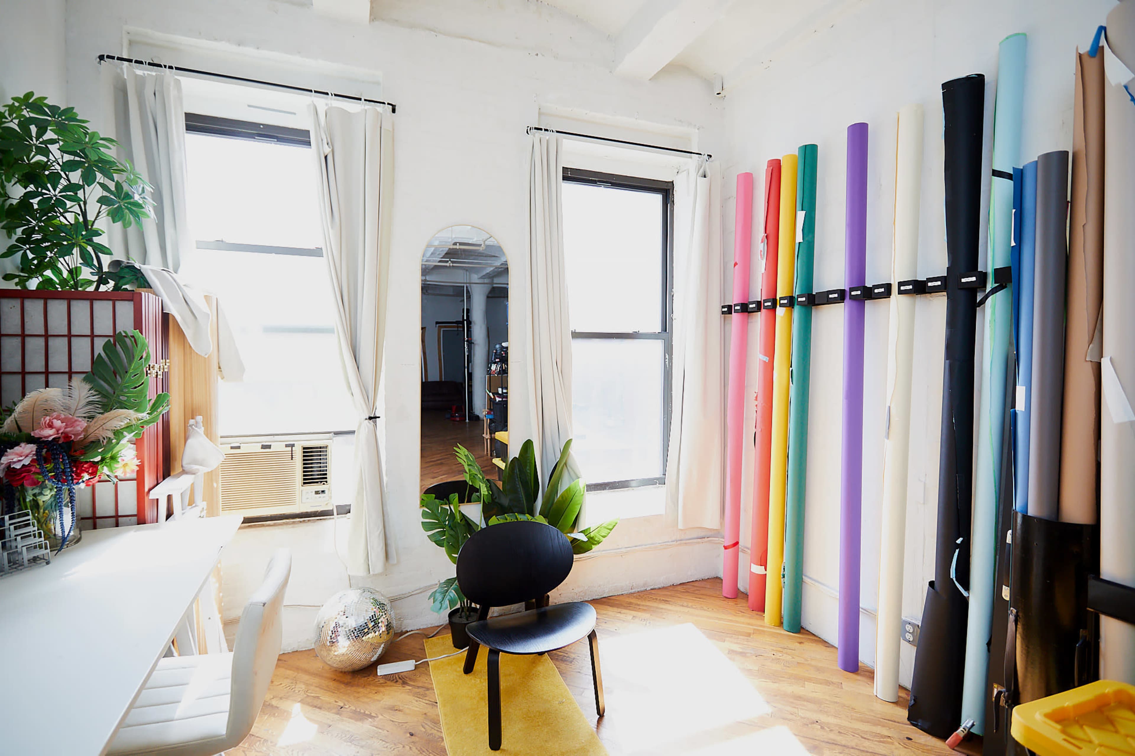 A bright studio space features a desk, plants, an air conditioning unit, and colorful rolls of paper neatly arranged against the wall.