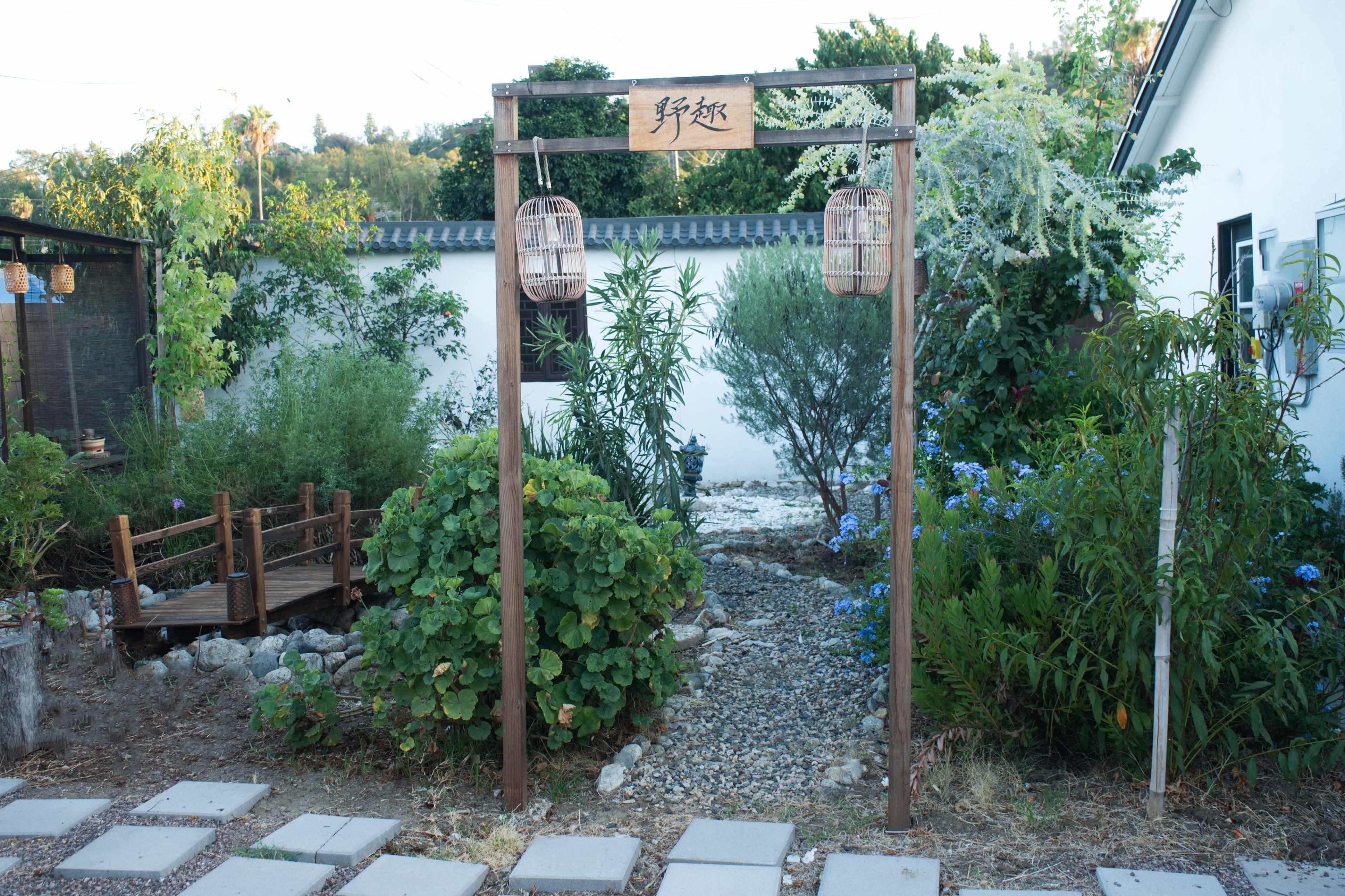 The image shows an entrance archway with a sign and lanterns, leading into a garden area with various plants and a wooden bridge.