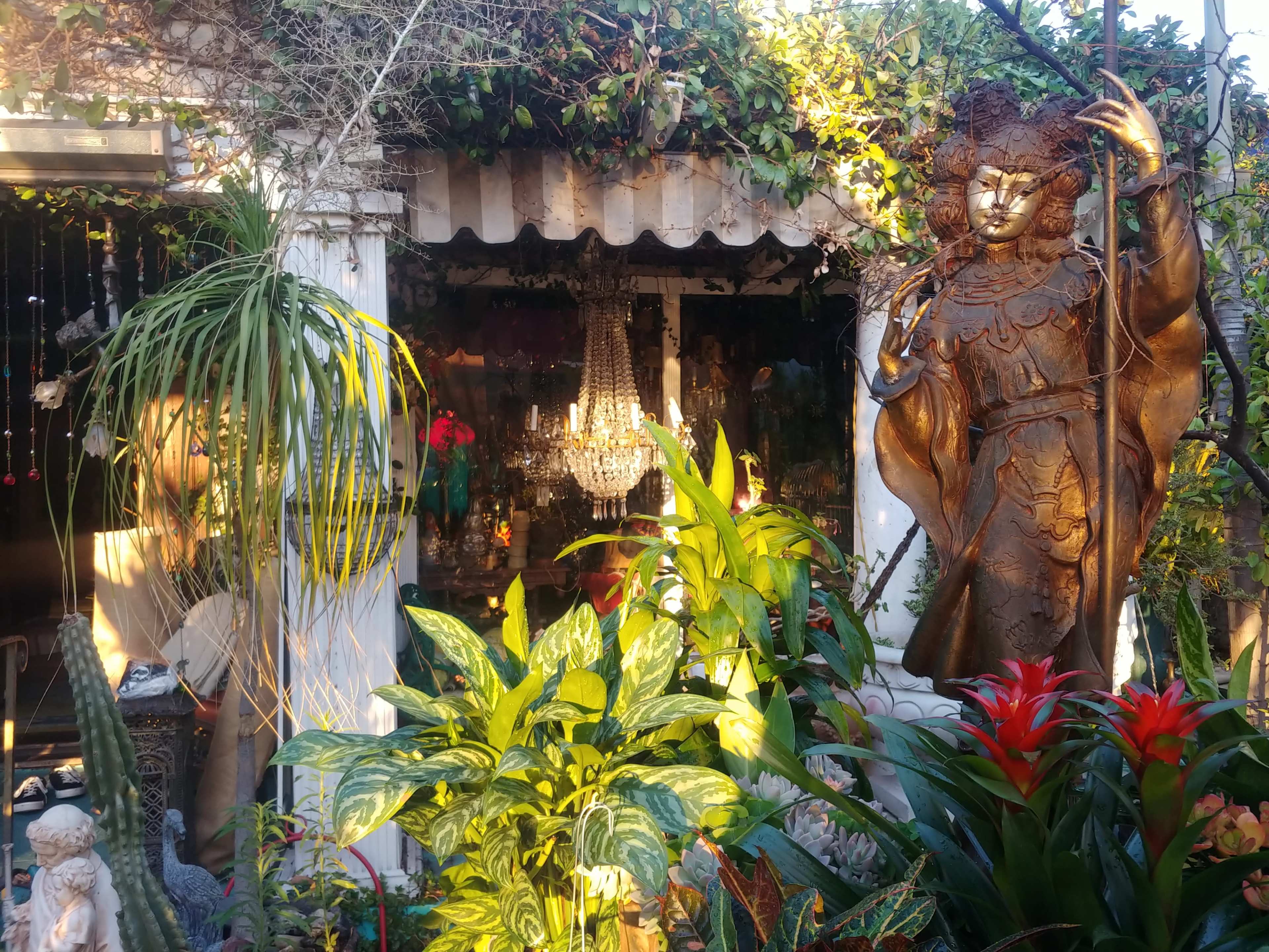 A large statue of a figure stands beside a lush garden filled with various plants, while a chandelier is visible in the background through an open doorway.