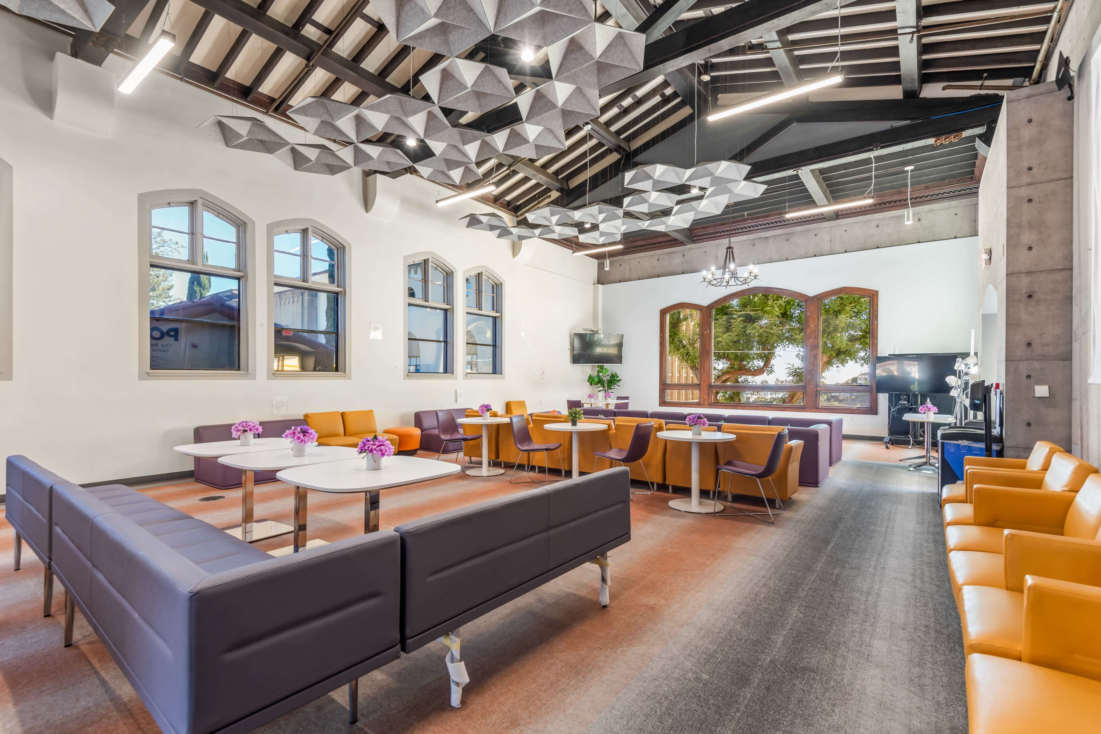The image shows a modern communal lounge area with a mix of purple and orange seating, large windows, and decorative ceiling panels.