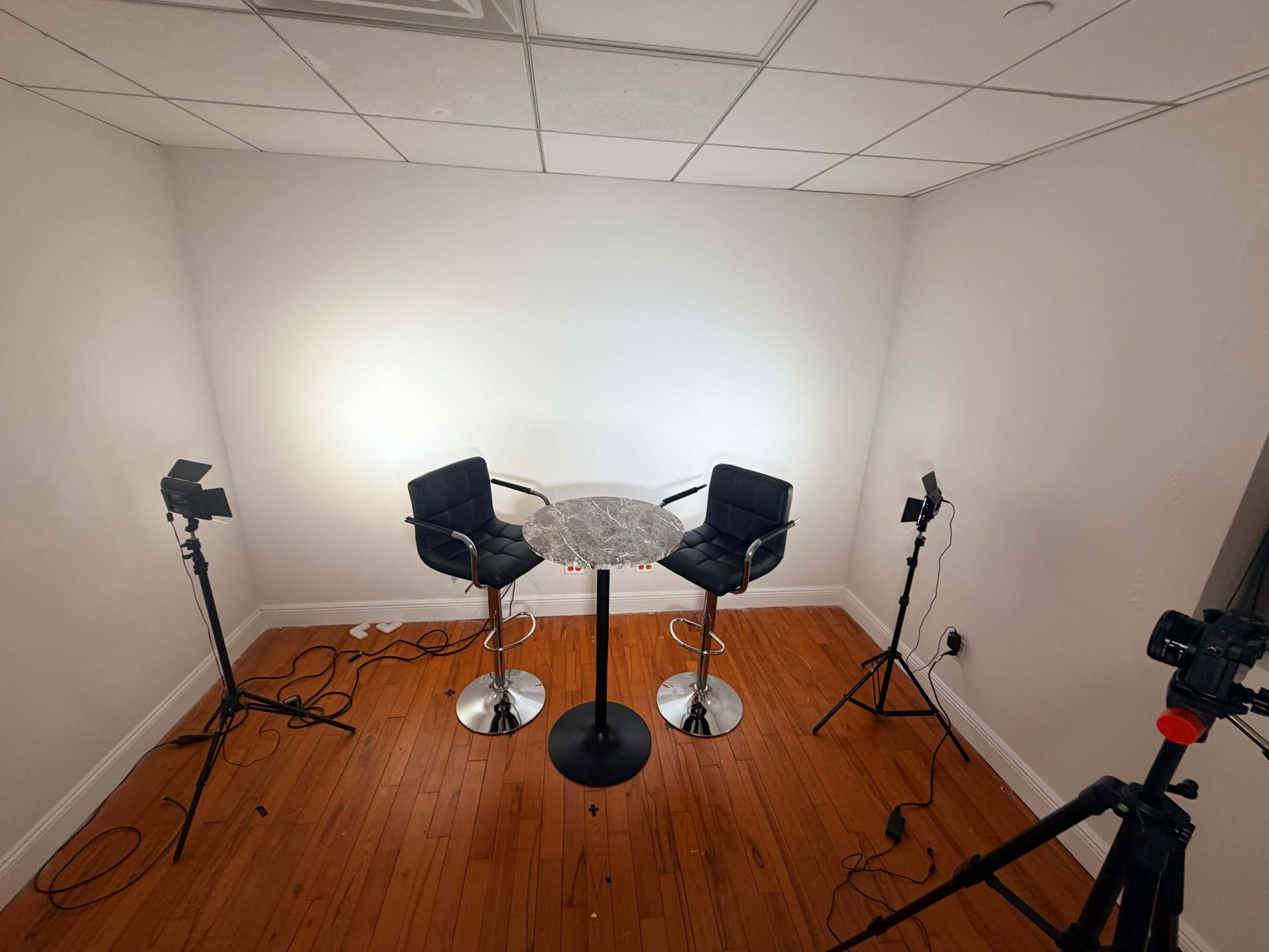The image shows a small, empty room with two black bar chairs and a round marble table, illuminated by three studio lights on stands.