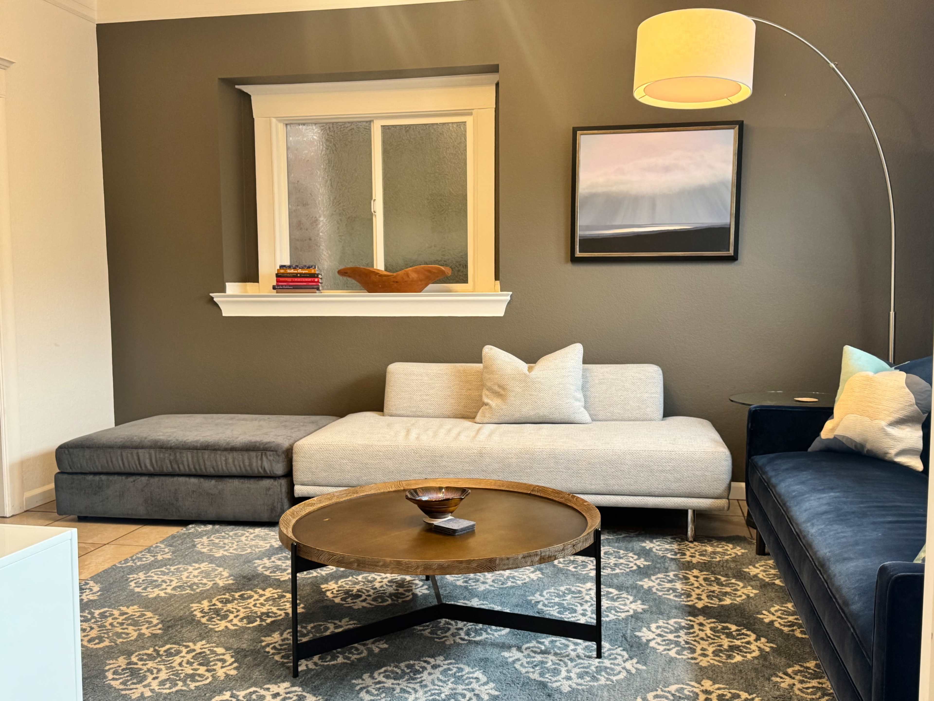 The image shows a modern living room with a gray sectional sofa, a round coffee table, and a floor lamp, complemented by a dark wall and decorative window.