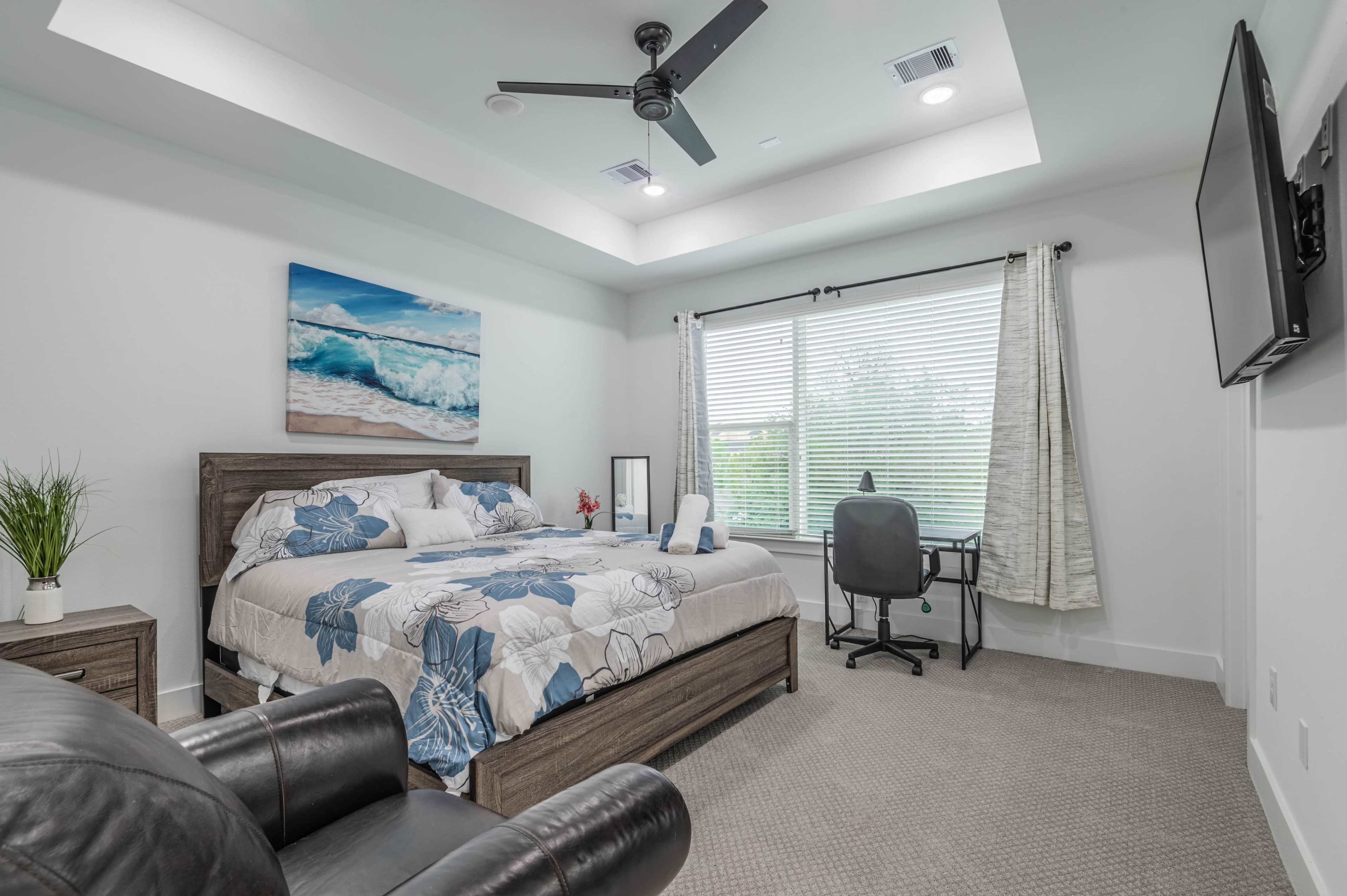A modern bedroom with a large bed, desk, and a window dressed in light curtains, featuring a white ceiling and a ceiling fan.